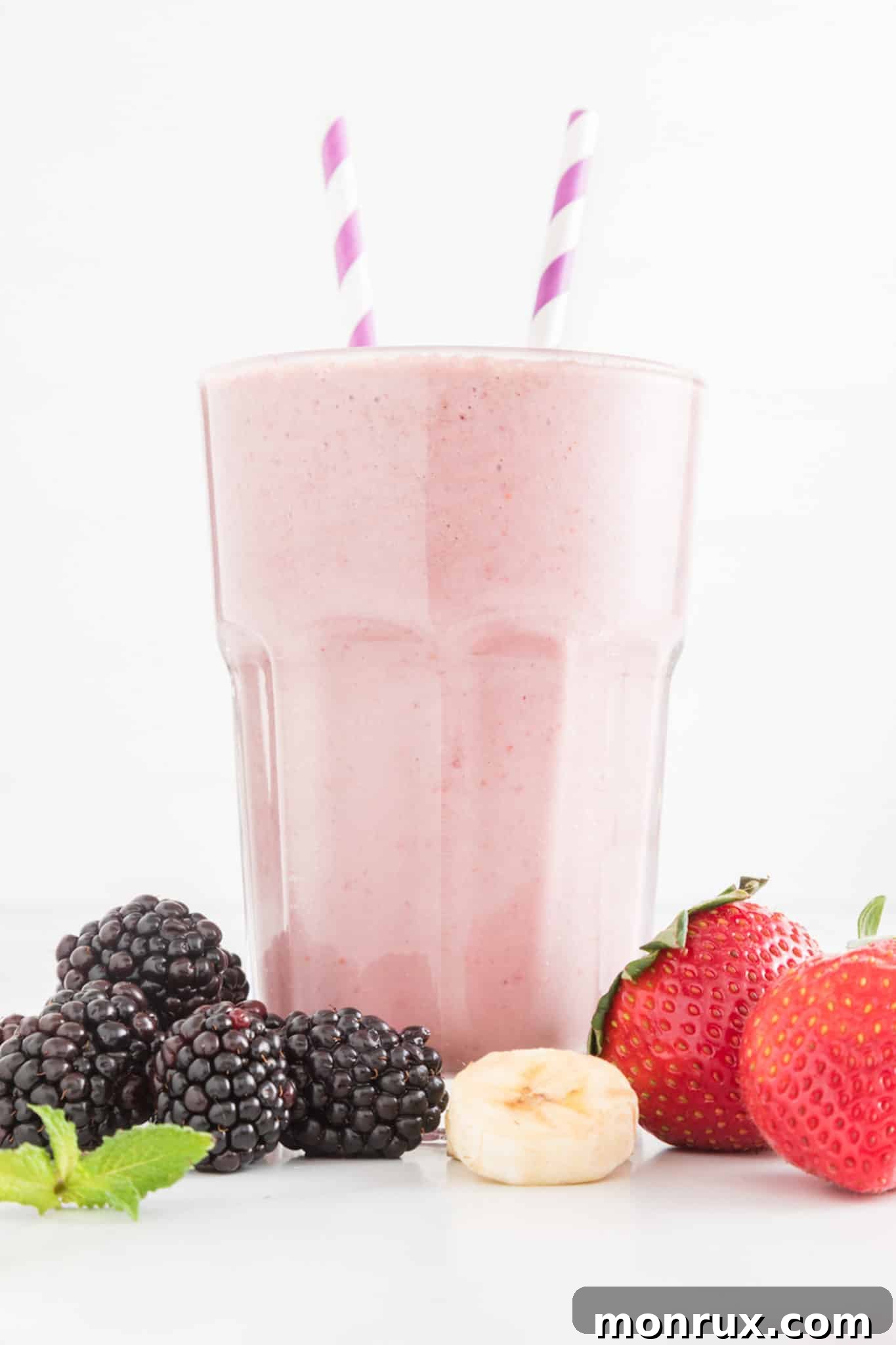 Two tall glasses of strawberry blackberry banana smoothie, garnished with fresh berries and two paper straws, placed on a light surface.