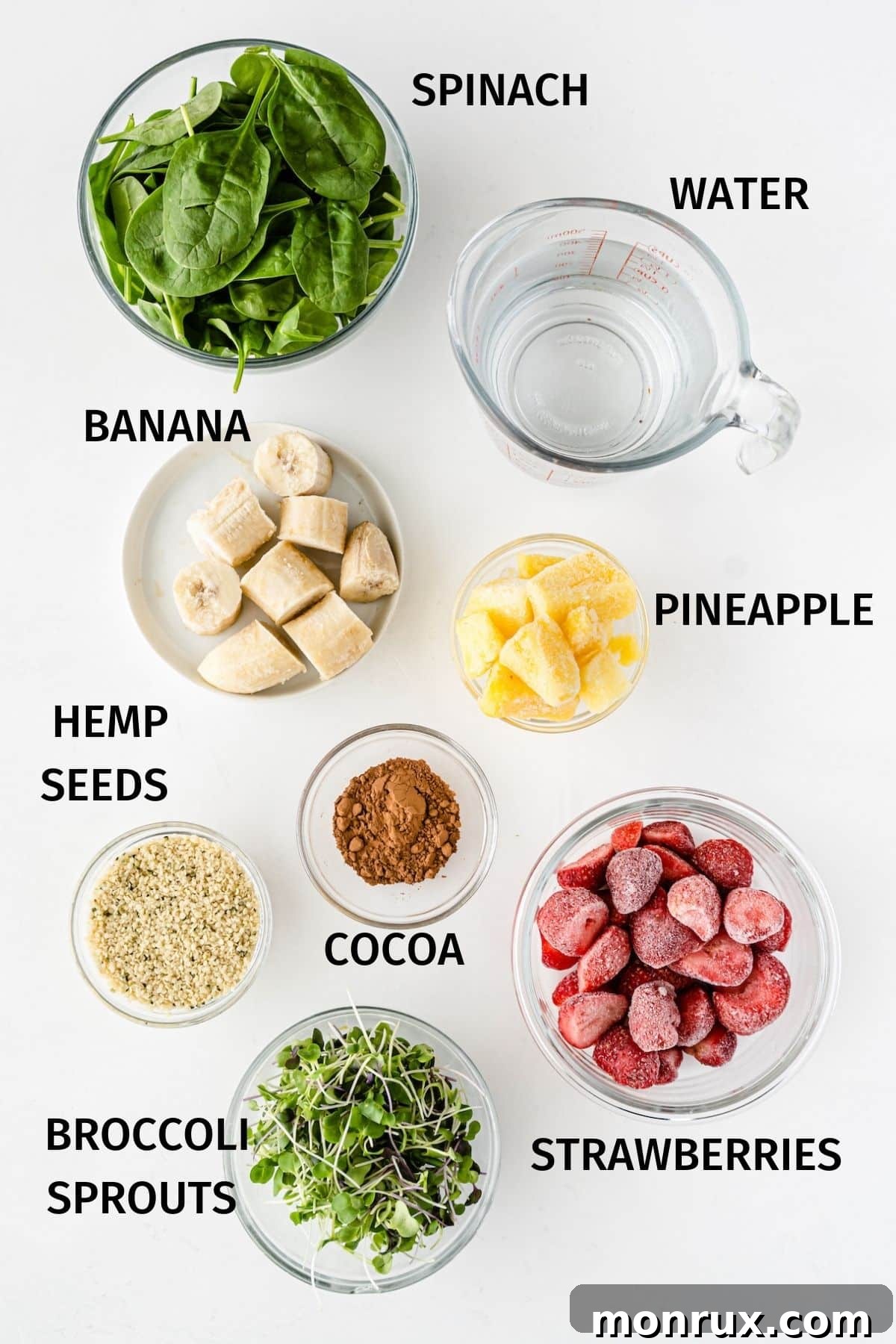 Fresh ingredients laid out on a wooden surface: filtered water, hemp seeds, baby spinach, broccoli sprouts, a frozen banana, frozen strawberries, frozen pineapple chunks, and cocoa powder.