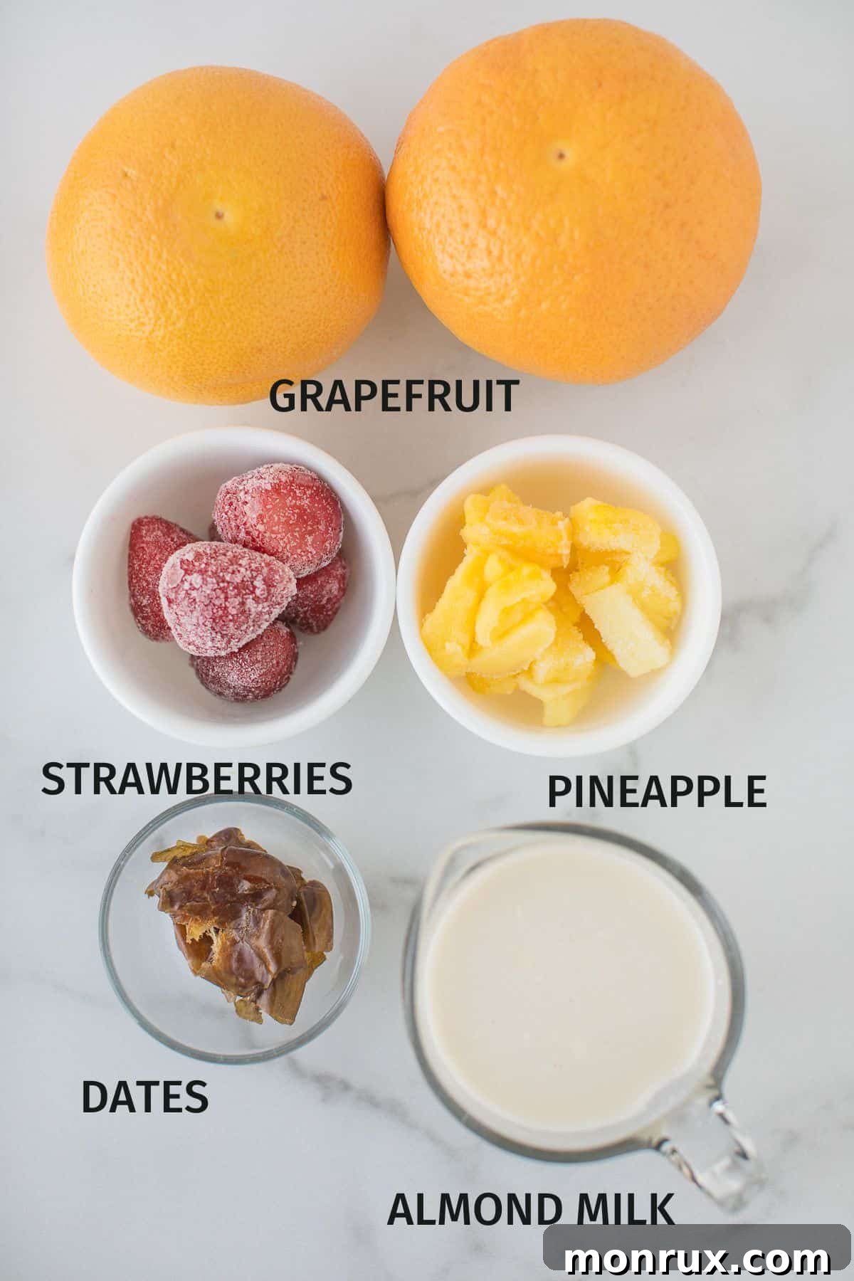 Labeled ingredients including grapefruit, frozen pineapple, frozen strawberries, almond milk, chia seeds, and dates arranged on a counter.