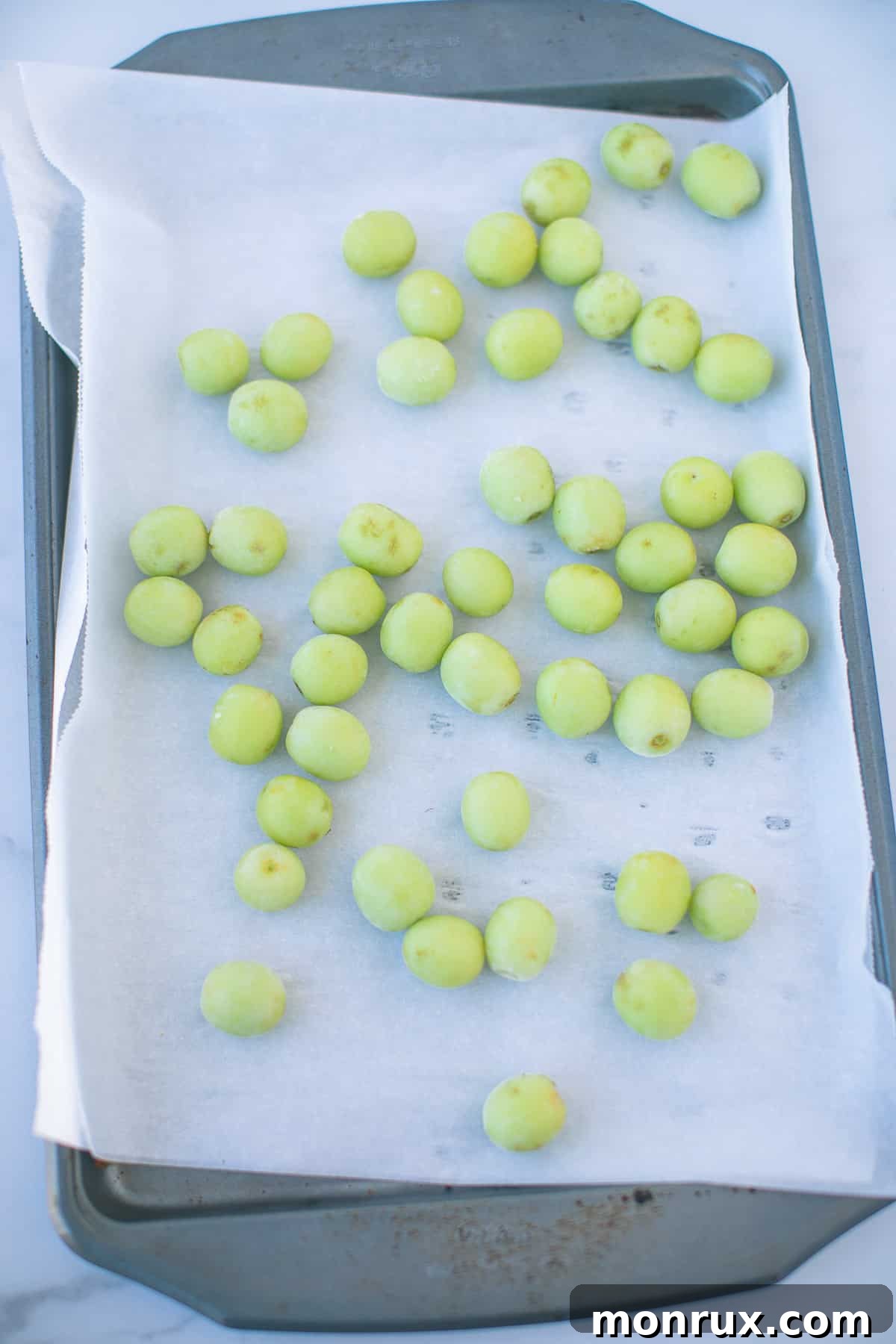 A baking sheet filled with individually frozen green grapes, showing a light frost, sits in a freezer.