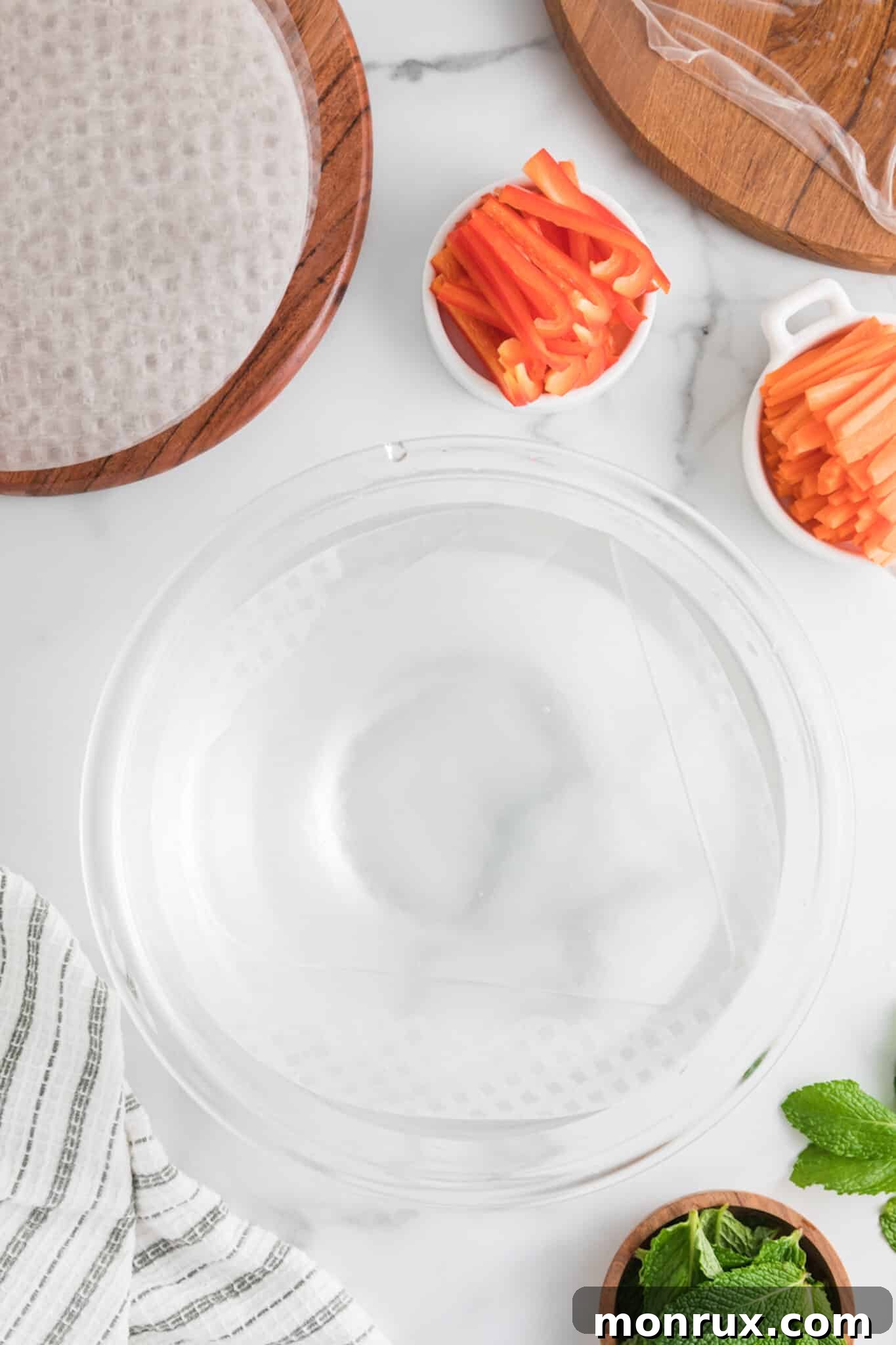 A close-up of a clear cutting board with a bowl of warm water and dry rice paper wrappers, ready for dipping.