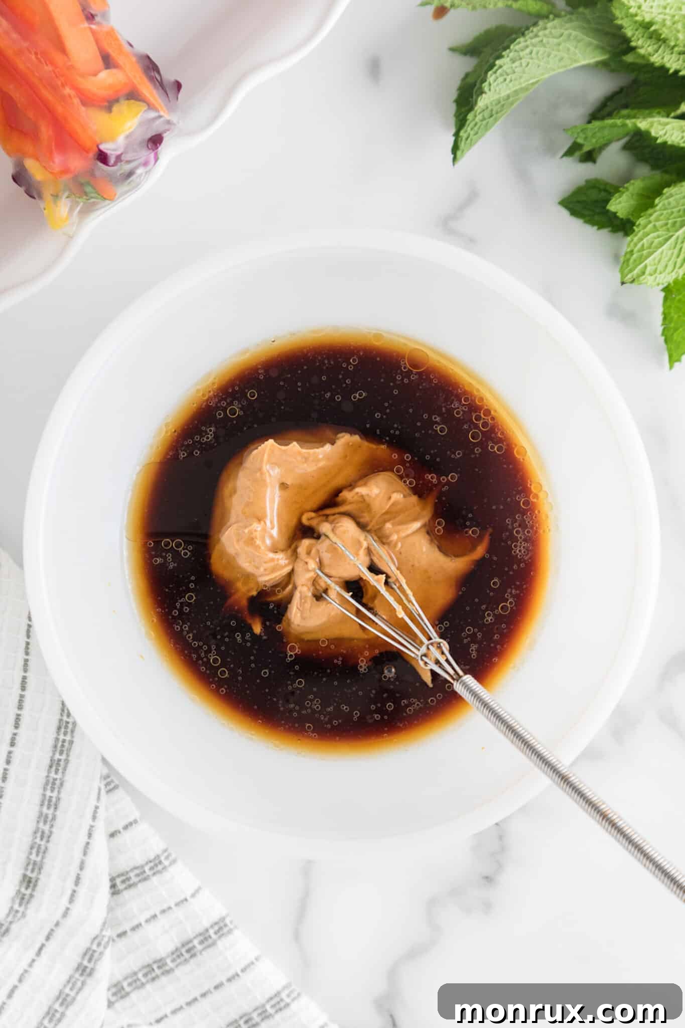 Ingredients for peanut dipping sauce, including peanut butter, coconut aminos, maple syrup, and sesame oil, in a bowl with a whisk.