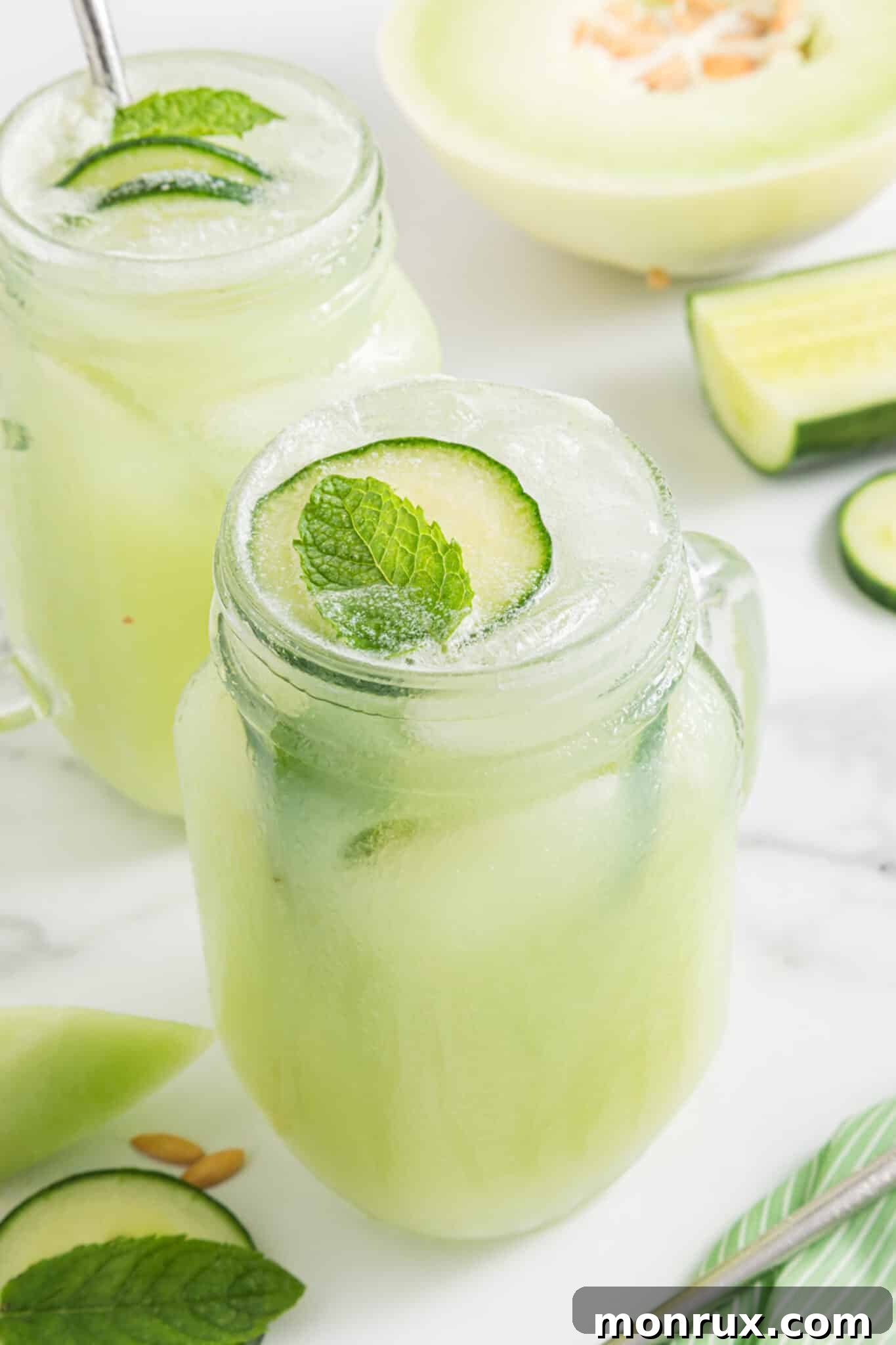 A handled mason jar filled with a bright green Honeydew Cucumber Smoothie, garnished with a cucumber slice and mint leaf, against a light background.