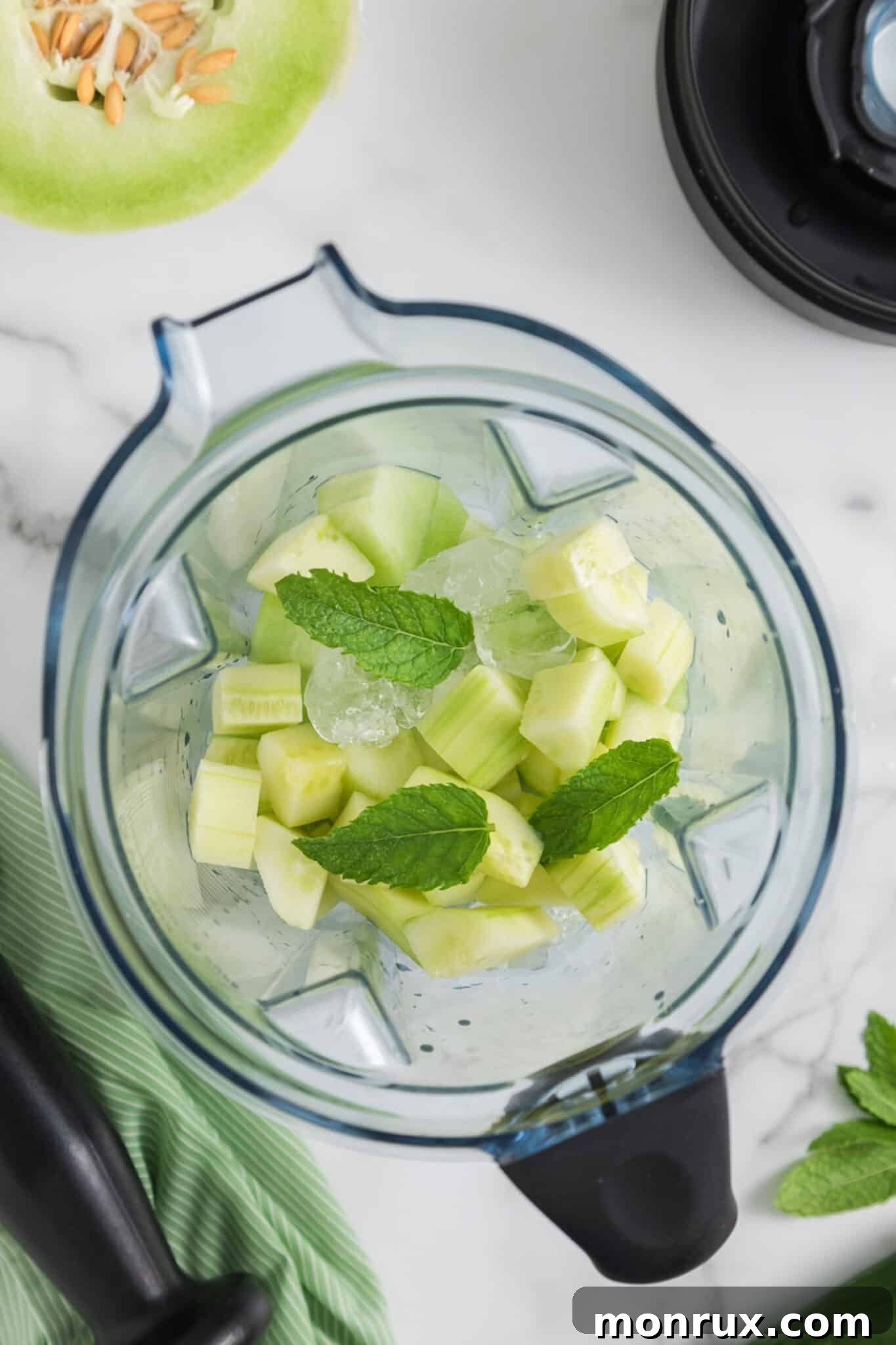 Chopped honeydew melon, cucumber, coconut milk, mint leaves, and ice placed in the jar of a high-speed blender, ready to be blended.