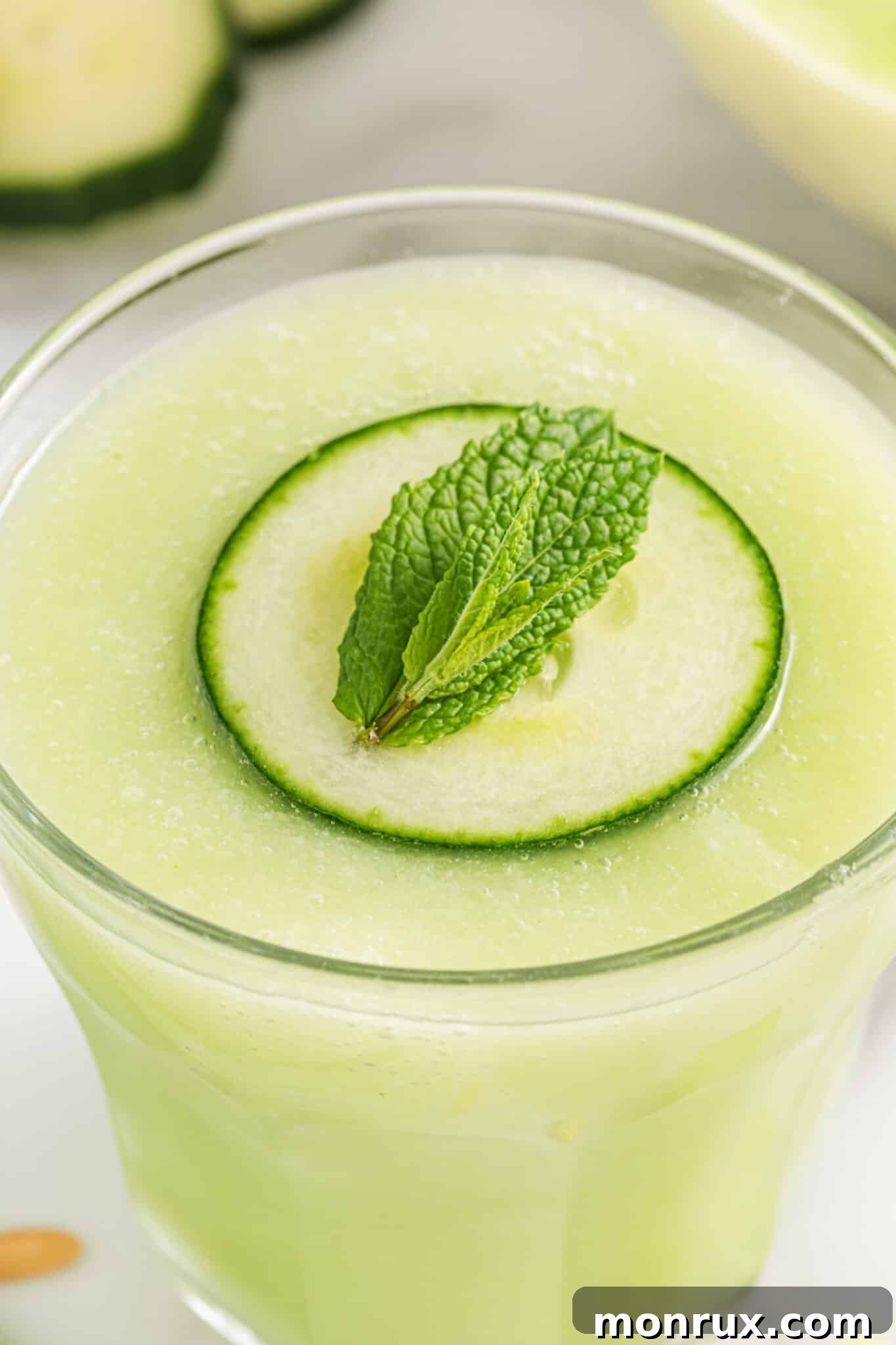 Closeup of a cucumber slice and a mint leaf elegantly floating on the surface of a clear glass filled with Honeydew Smoothie.