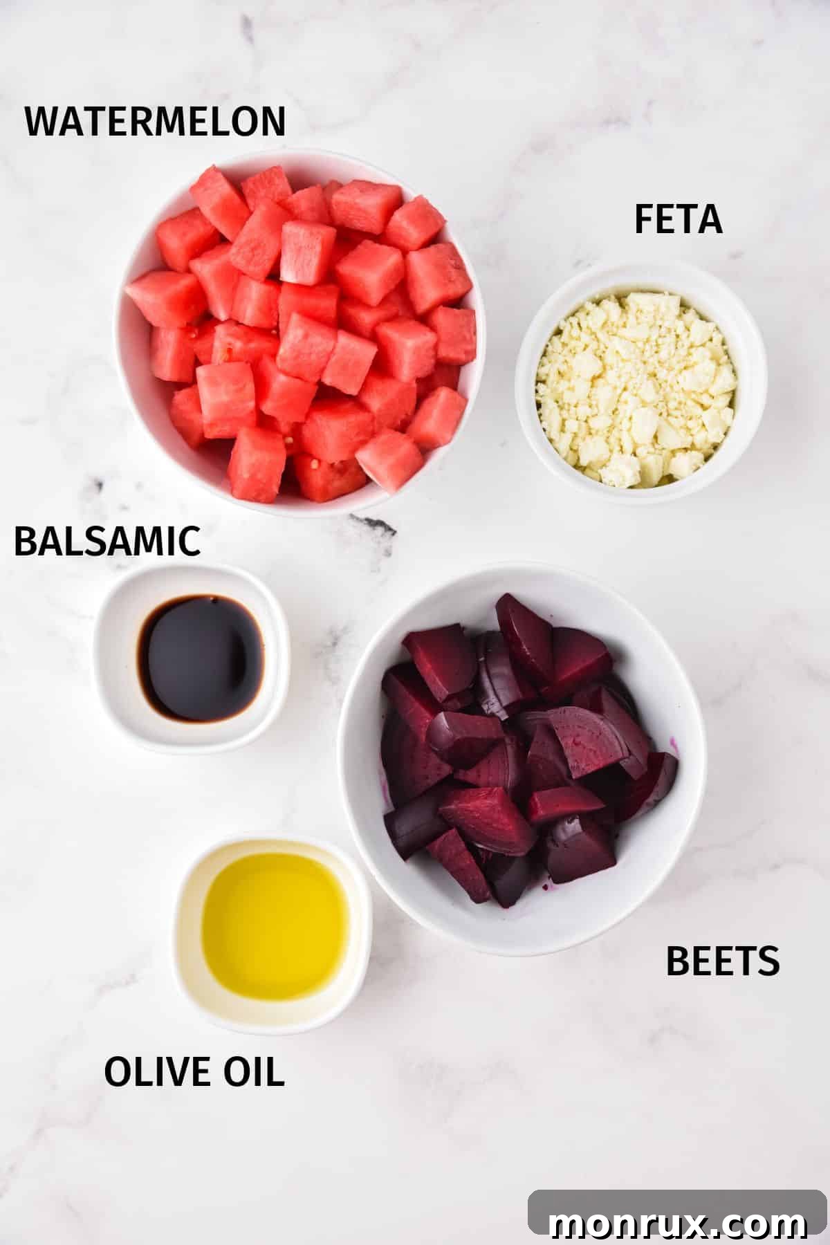 Small white bowls filled with the ingredients for watermelon beet salad: cubed watermelon, roasted beets, crumbled feta, olive oil, and balsamic vinegar.