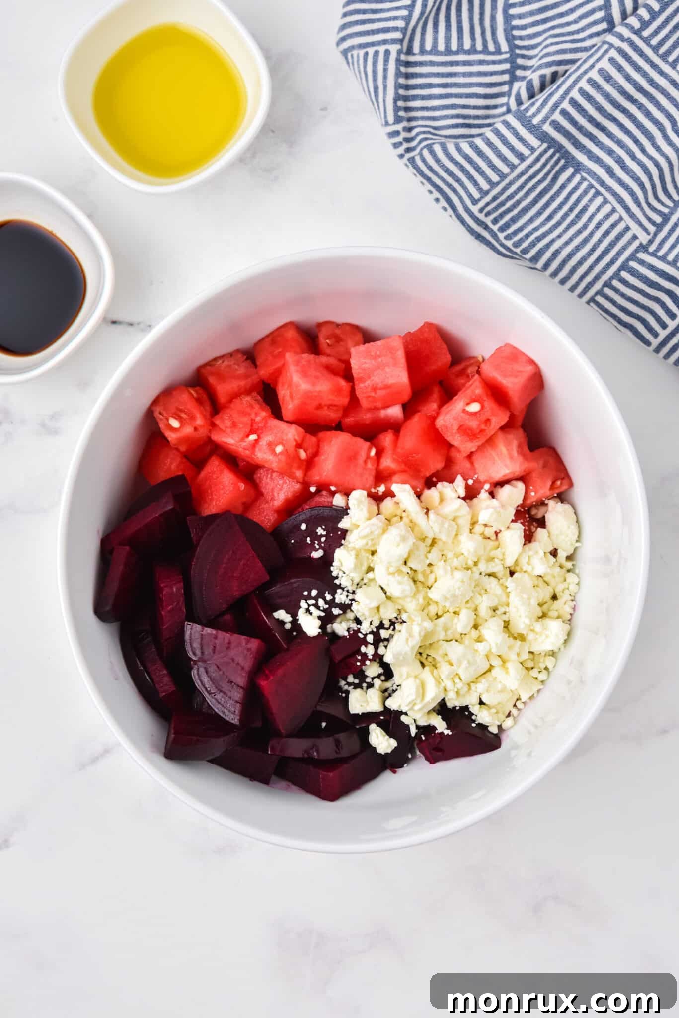 Cubed roasted beets, fresh watermelon pieces, and crumbled feta cheese are combined in a large white serving bowl, ready for dressing.