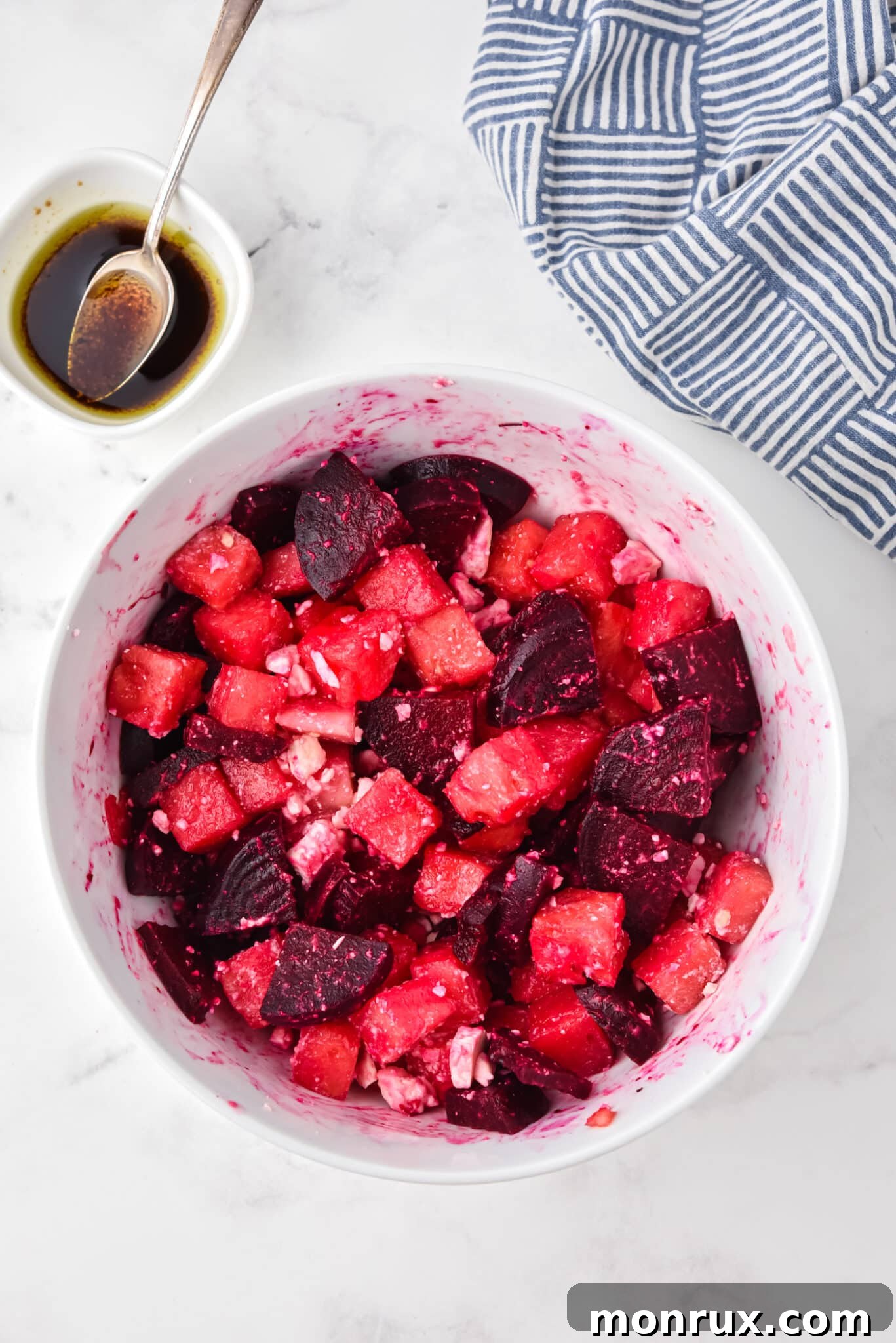 A hand drizzling and stirring balsamic vinaigrette dressing into a bowl of vibrant watermelon beet salad, coating the colorful cubes of fruit and vegetable.