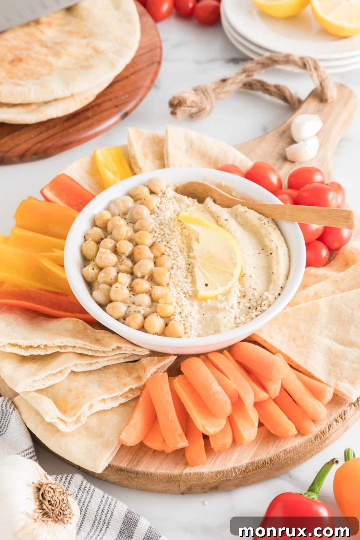 A beautiful wooden platter laden with colorful sliced vegetables like carrots, cucumbers, and bell peppers, along with quartered pita bread arranged around a small, elegant bowl of homemade oil-free hummus. The hummus is garnished lightly.