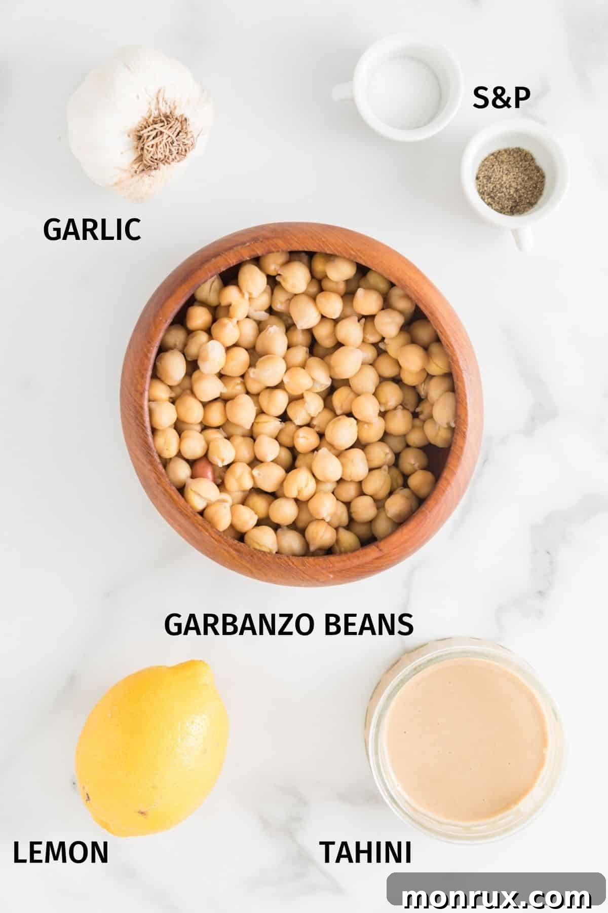 Small bowls containing the key ingredients for oil-free hummus: chickpeas, tahini, fresh lemon halves, and a garlic clove, neatly arranged on a white surface.