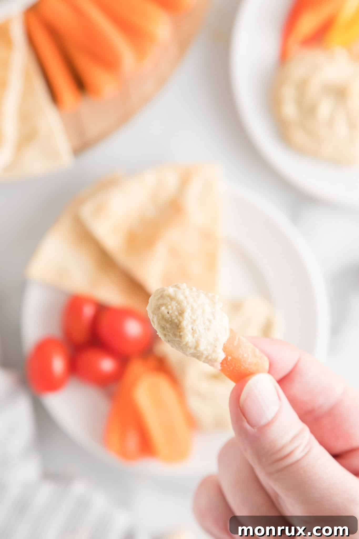 A close-up shot of a hand holding a vibrant baby carrot, freshly dipped into a bowl of homemade, creamy oil-free hummus.