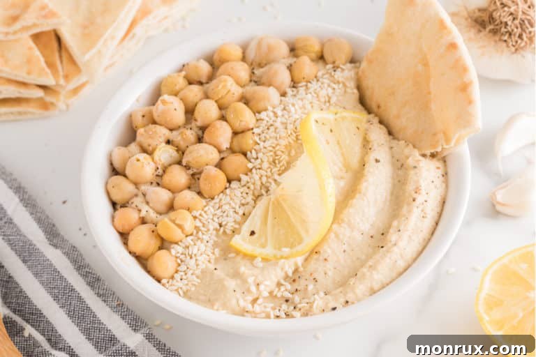 A white ceramic bowl filled with creamy homemade oil-free hummus, artfully garnished with a sprinkle of paprika, whole chickpeas, and a drizzle of (optional) extra virgin olive oil.