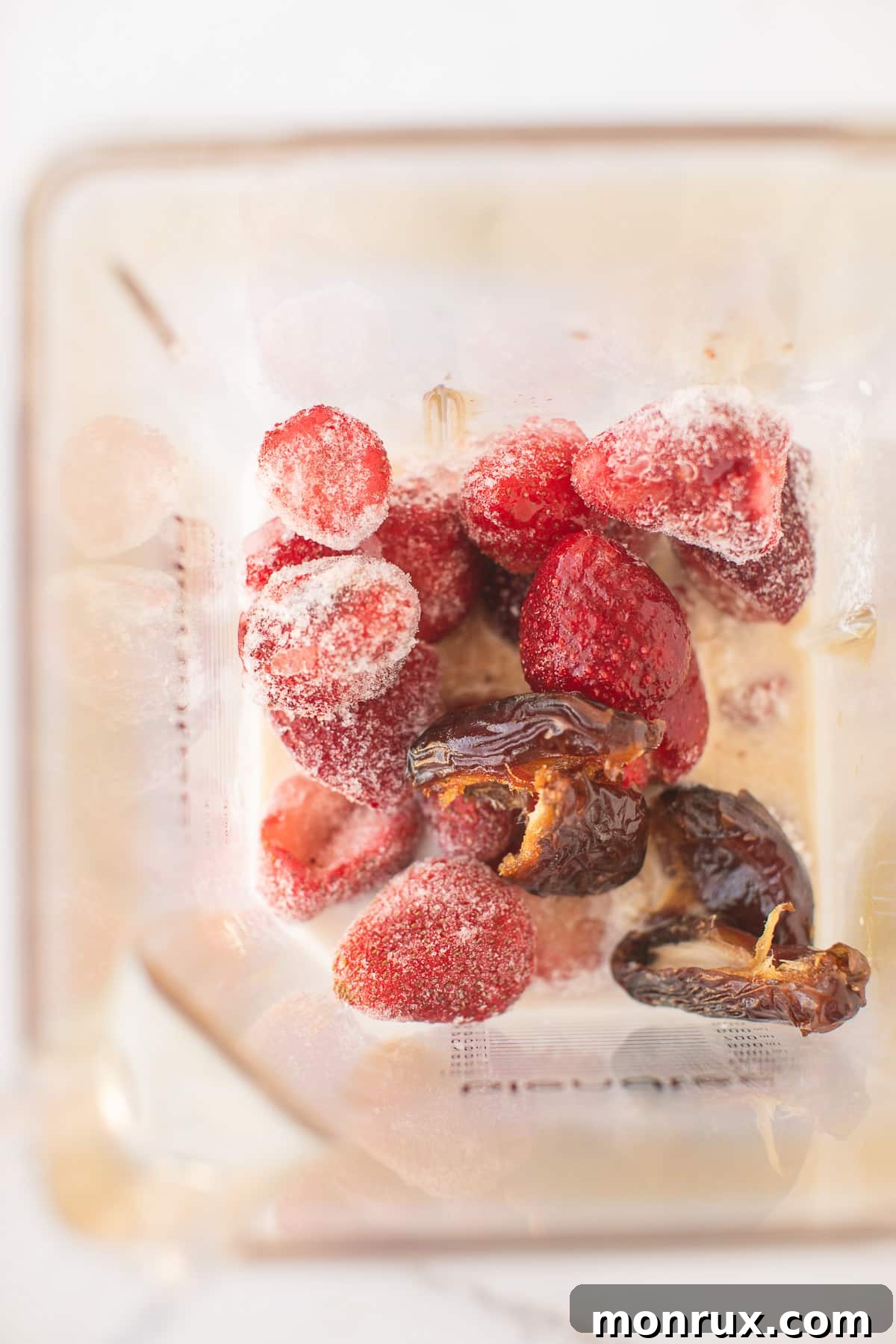 A high-speed blender pitcher, partially filled with the vibrant ingredients of a strawberry milkshake—coconut milk, frozen strawberries, and dates—poised for blending.