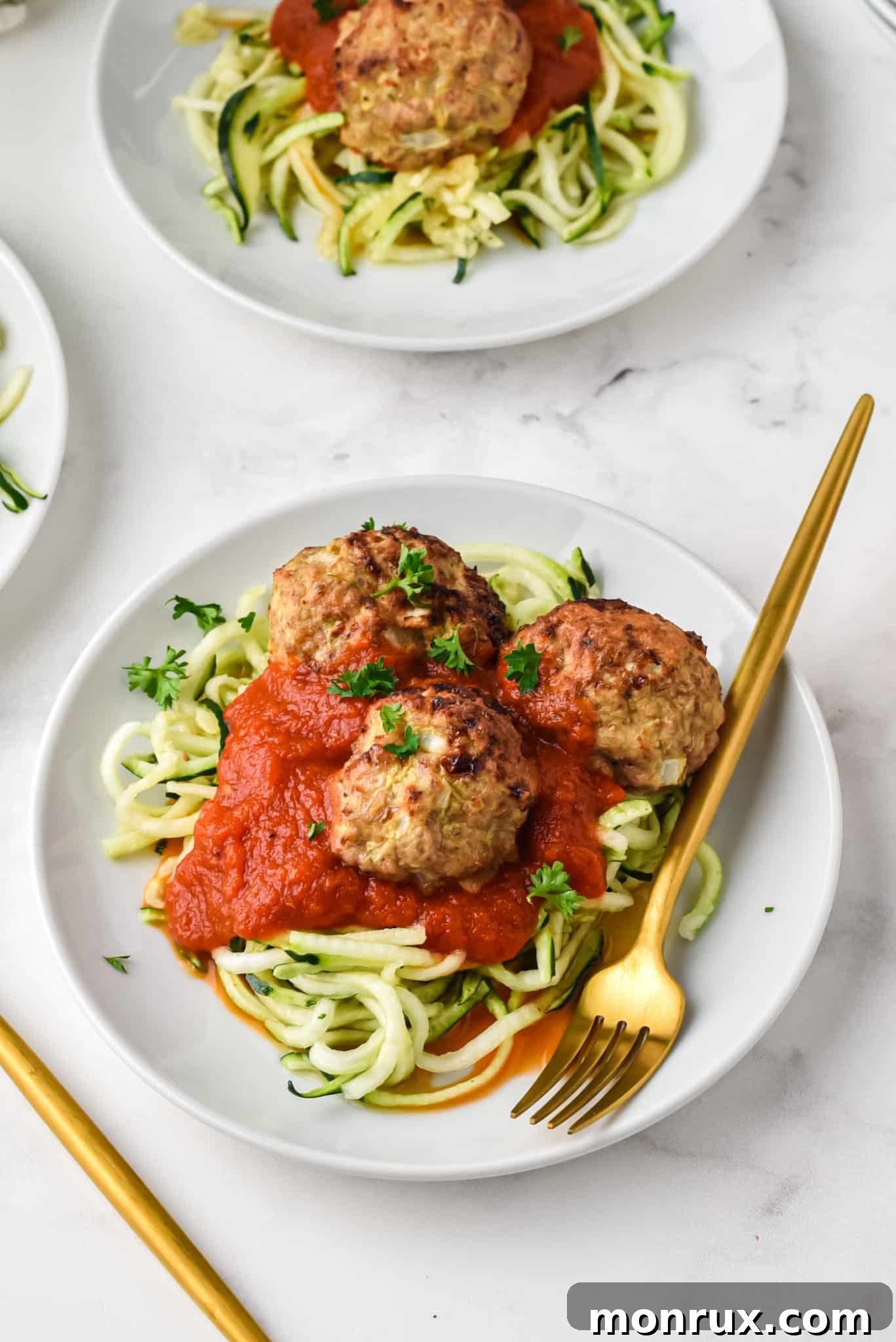 Two plates of zoodles topped with rich red sauce and tender turkey meatballs, garnished with fresh herbs.