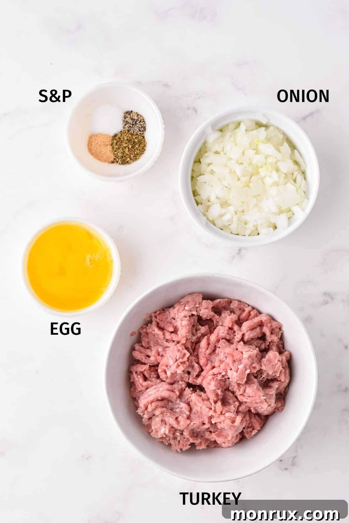 Ingredients for air fryer turkey meatballs in small bowls on a white surface, including ground turkey, egg, onion, and spices.