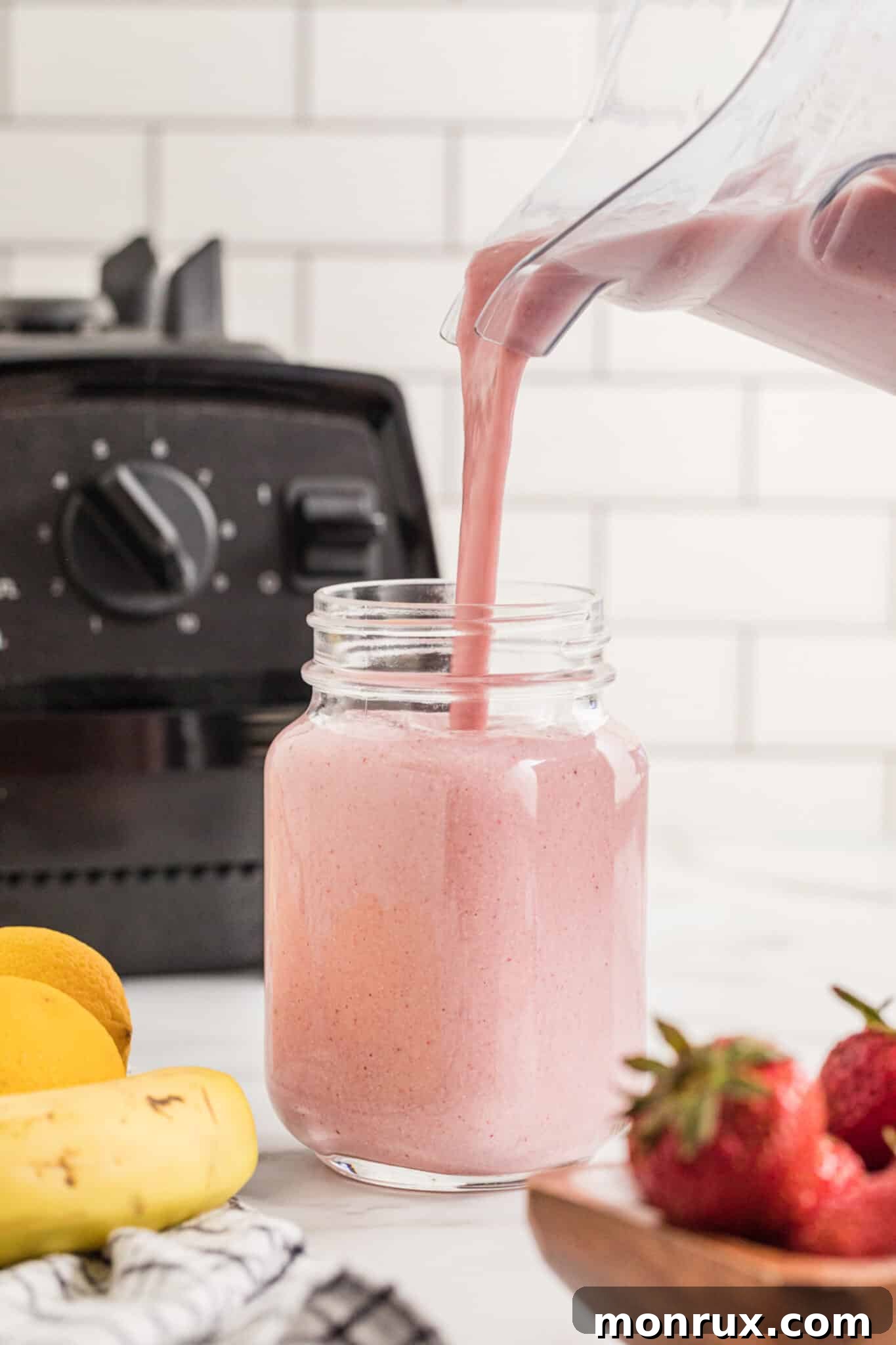 A rich, pink Strawberry Cheesecake Smoothie gracefully being poured from a high-speed blender into a rustic mason jar.
