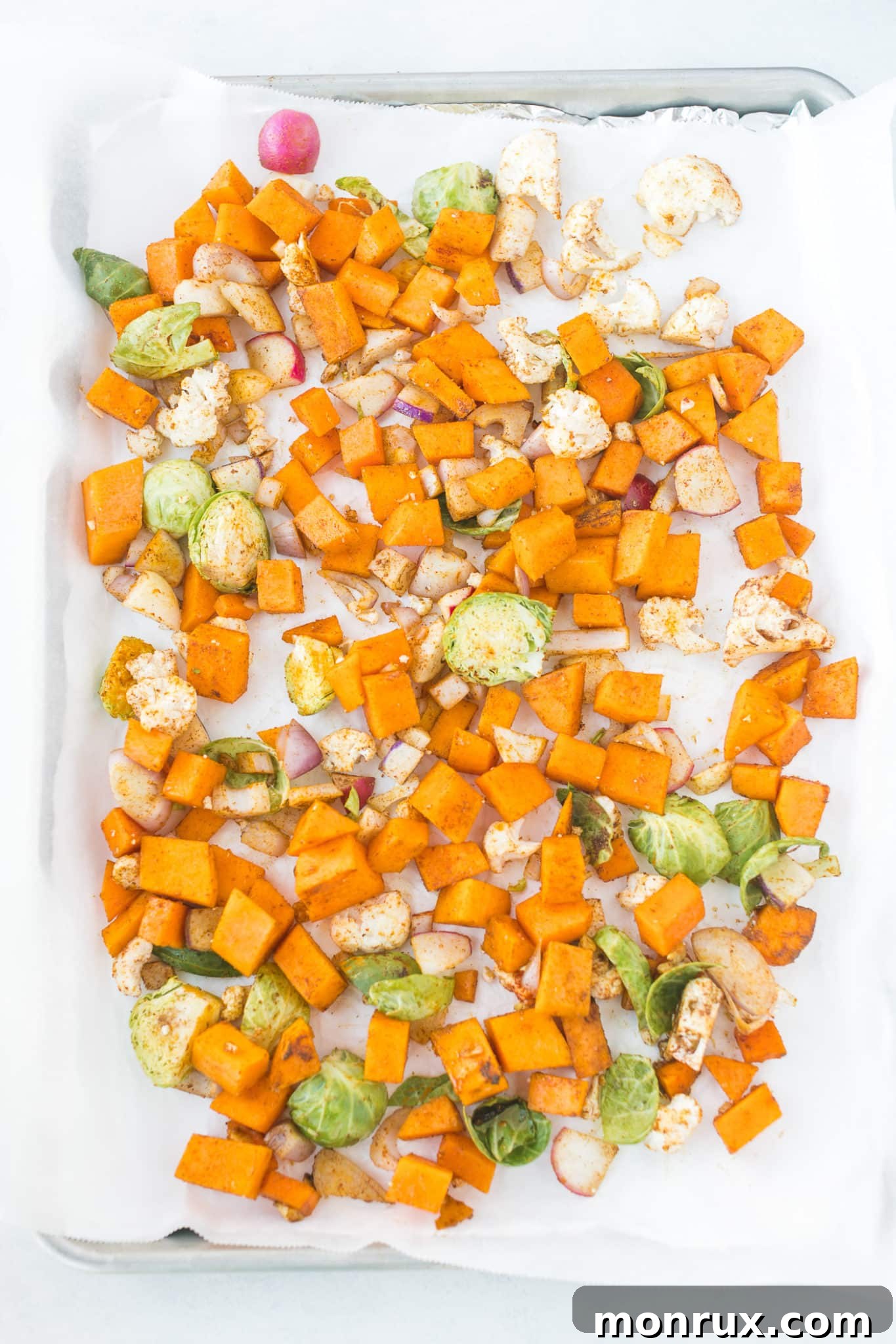 Seasoned mixed vegetables, including butternut squash and Brussels sprouts, spread out on a parchment paper-lined baking sheet, ready to be roasted in the oven.