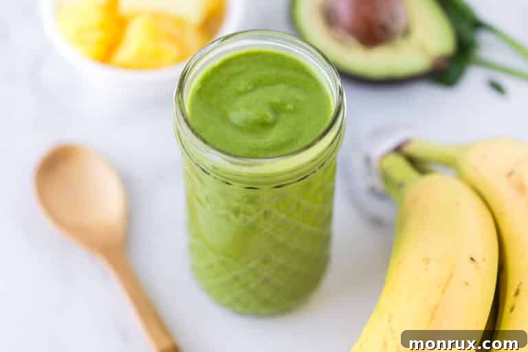 detox smoothie in a glass surrounded by a spoon, bananas, avocado, and pineapple