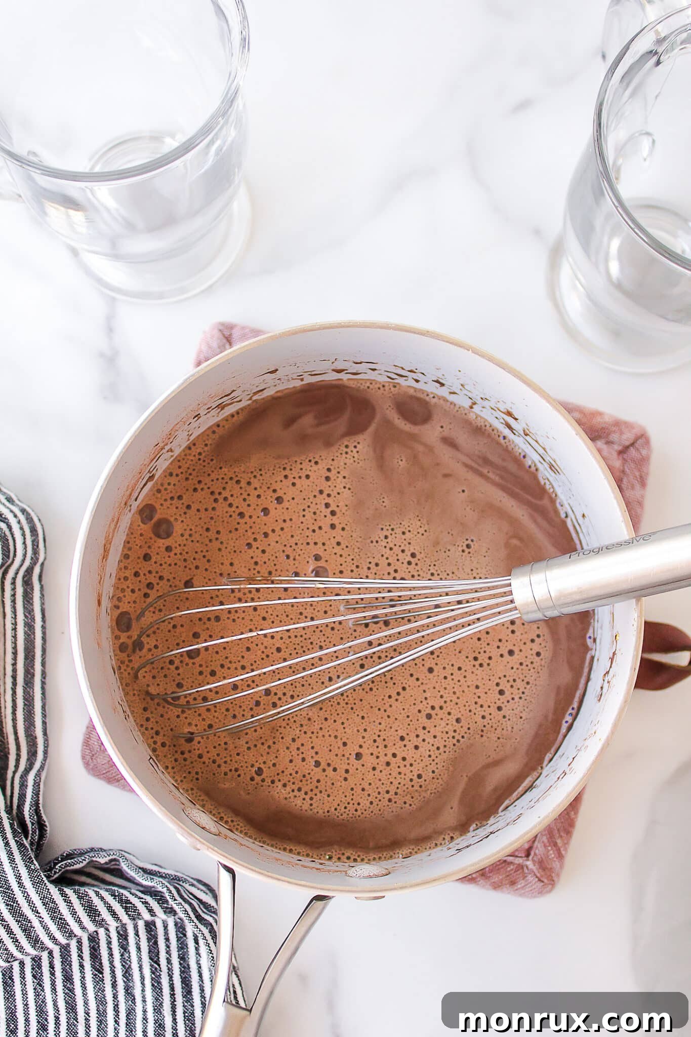 A whisk actively mixing cocoa powder into the warming milk and other ingredients in a saucepan, ensuring a smooth, lump-free consistency for the hot chocolate.