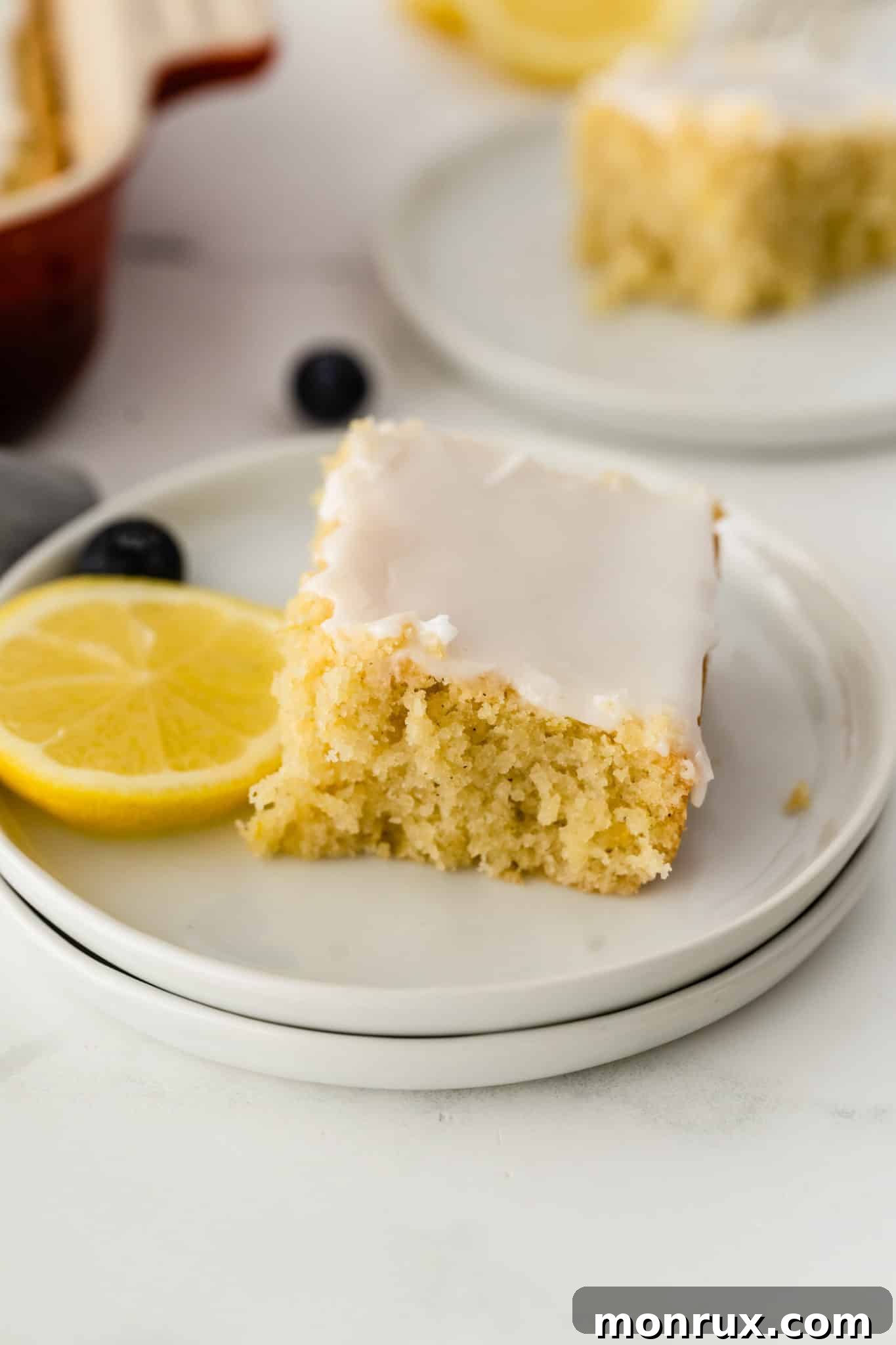 A beautifully sliced gluten-free lemon cake with a generous icing drizzle, garnished with fresh lemon zest, served on a white plate.