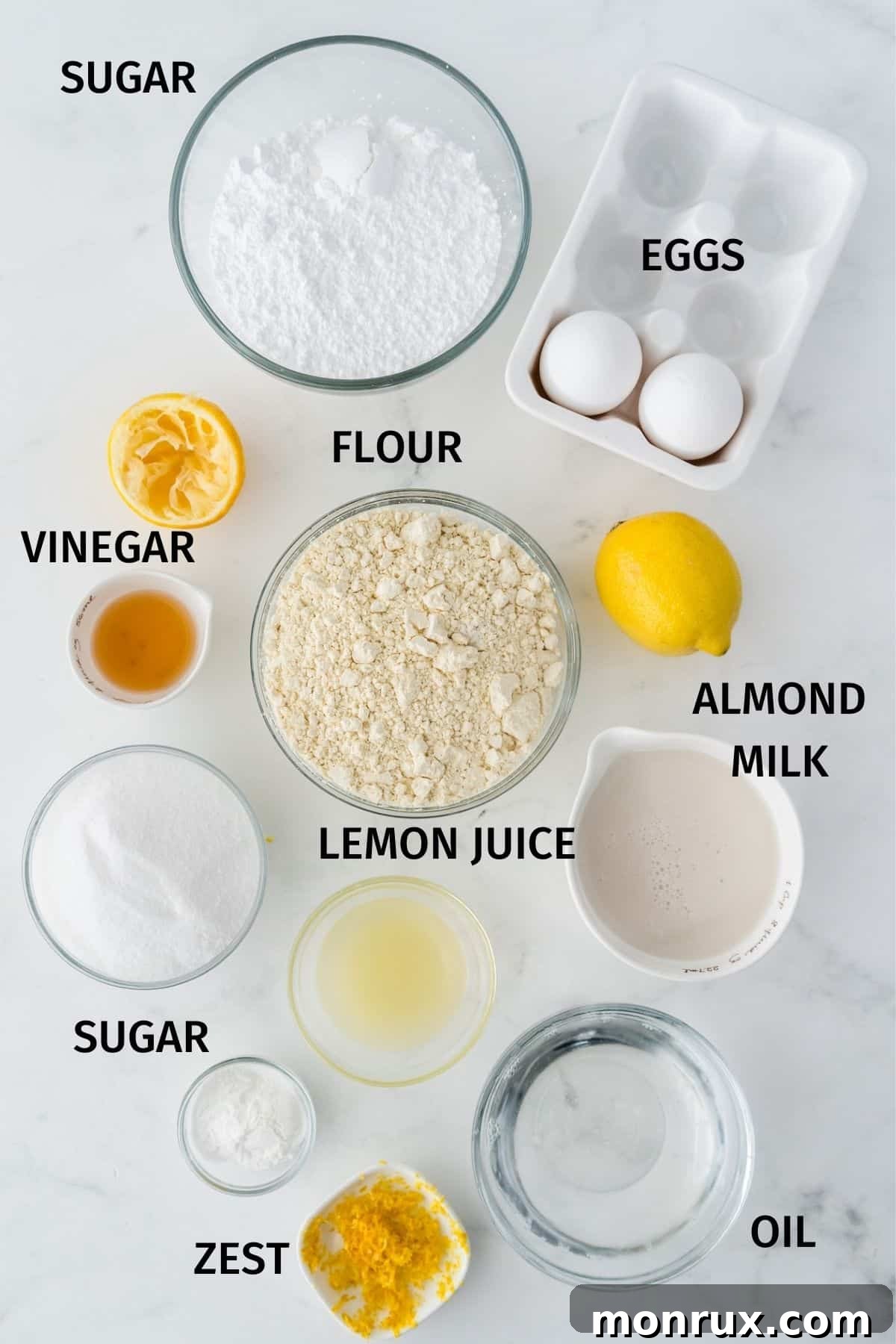 Collection of labeled ingredients including gluten-free flour, almond milk, lemon, eggs, coconut oil, and powdered sugar, laid out on a white surface for a gluten-free lemon drizzle cake recipe.