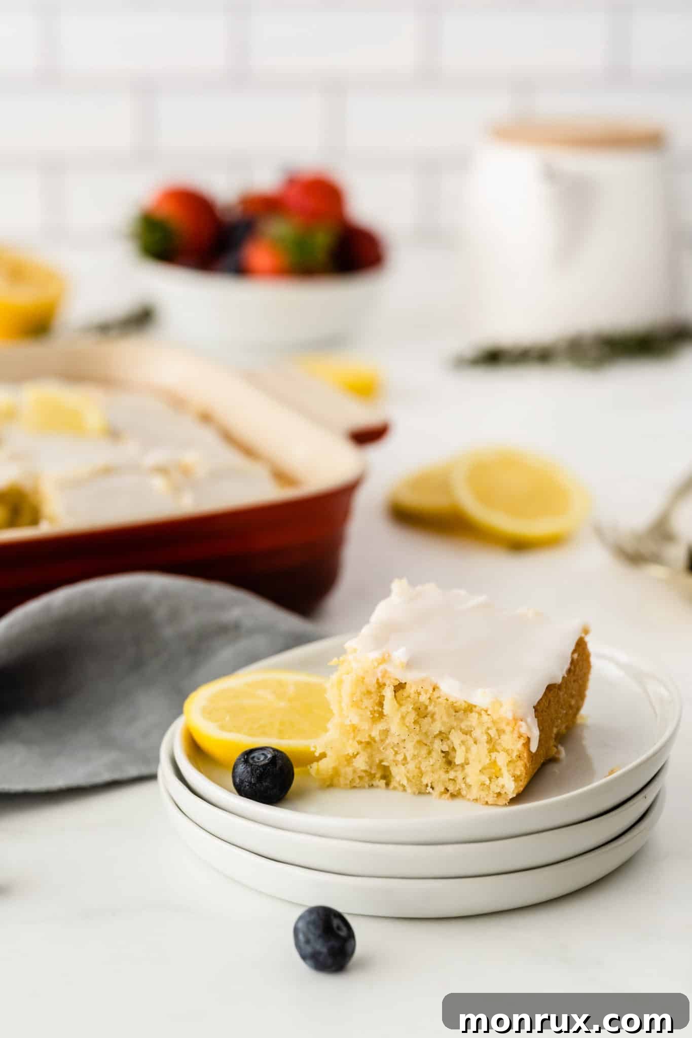 A close-up slice of gluten-free lemon drizzle cake, showing its moist texture and shiny glaze, presented on a stack of white plates.