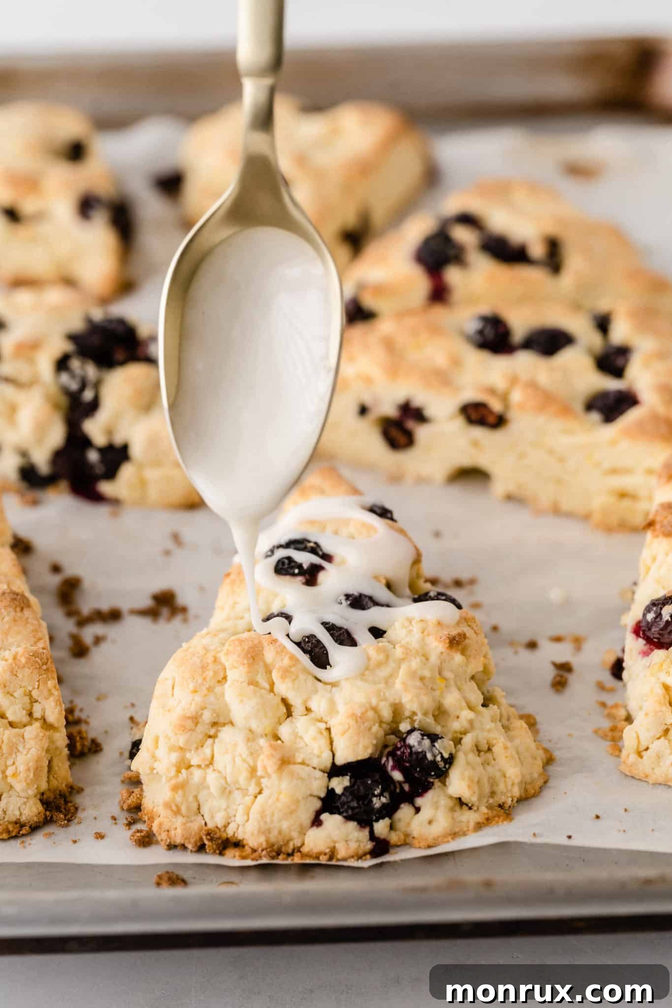A close-up shot of a spoon expertly drizzling a sweet lemon glaze over a batch of freshly baked gluten-free blueberry scones, creating a tempting finish.