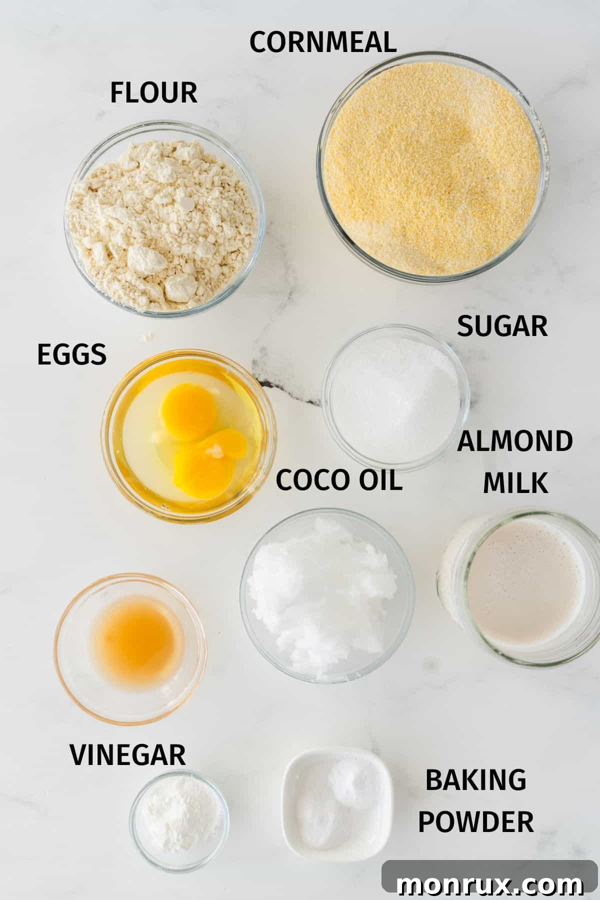 A collection of ingredients laid out for making gluten-free and dairy-free cornbread, including flour, cornmeal, almond milk, and eggs.