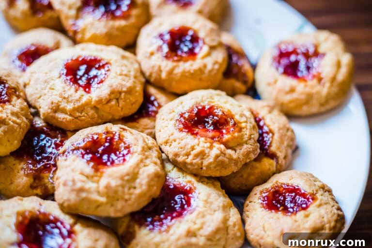 A plate piled high with golden gluten-free thumbprint cookies, each showcasing a vibrant, glistening jam center.