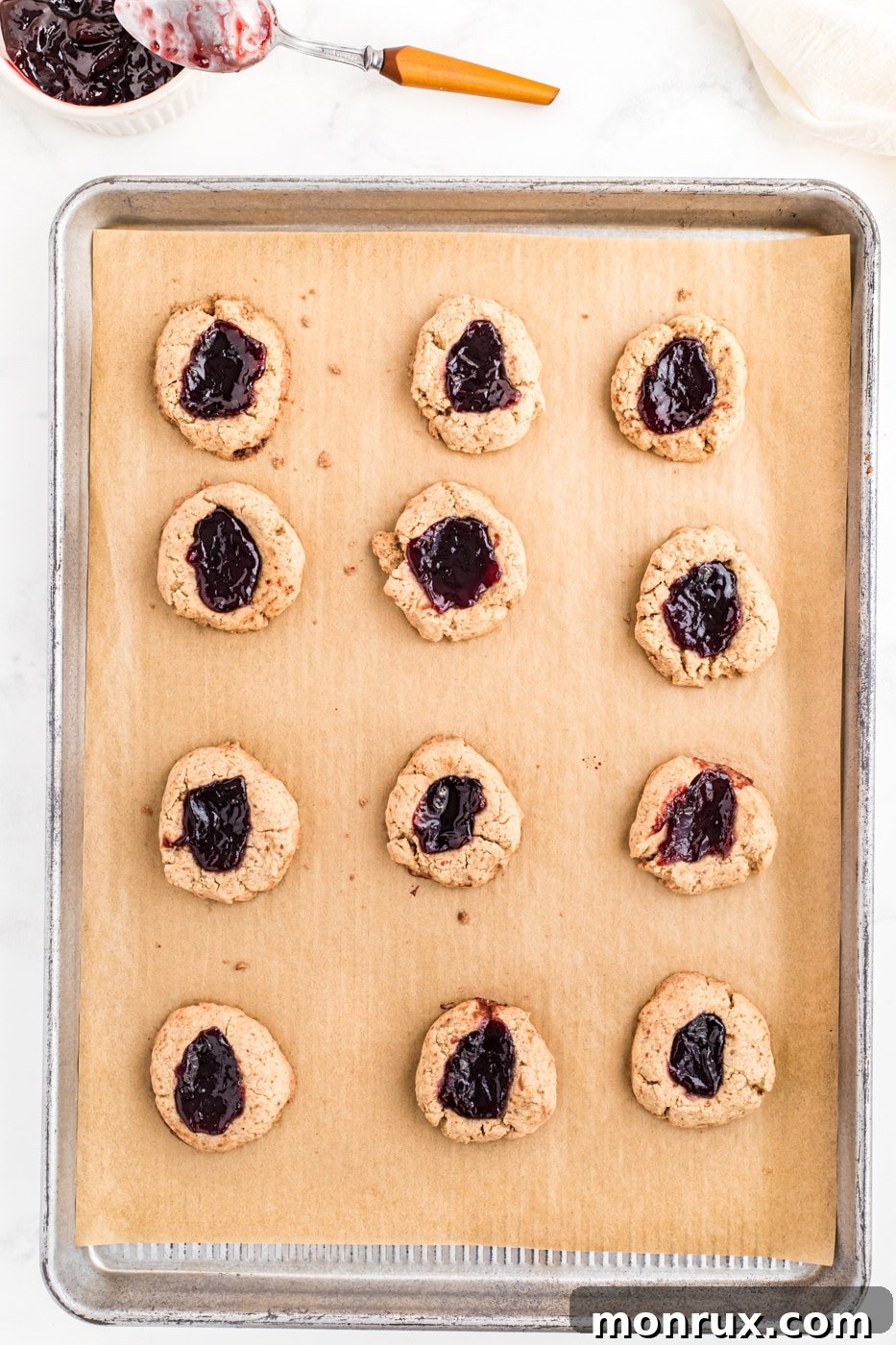 Warm, jam-filled thumbprint cookies resting on a baking sheet, fresh out of the oven, with their vibrant fruit centers glistening.