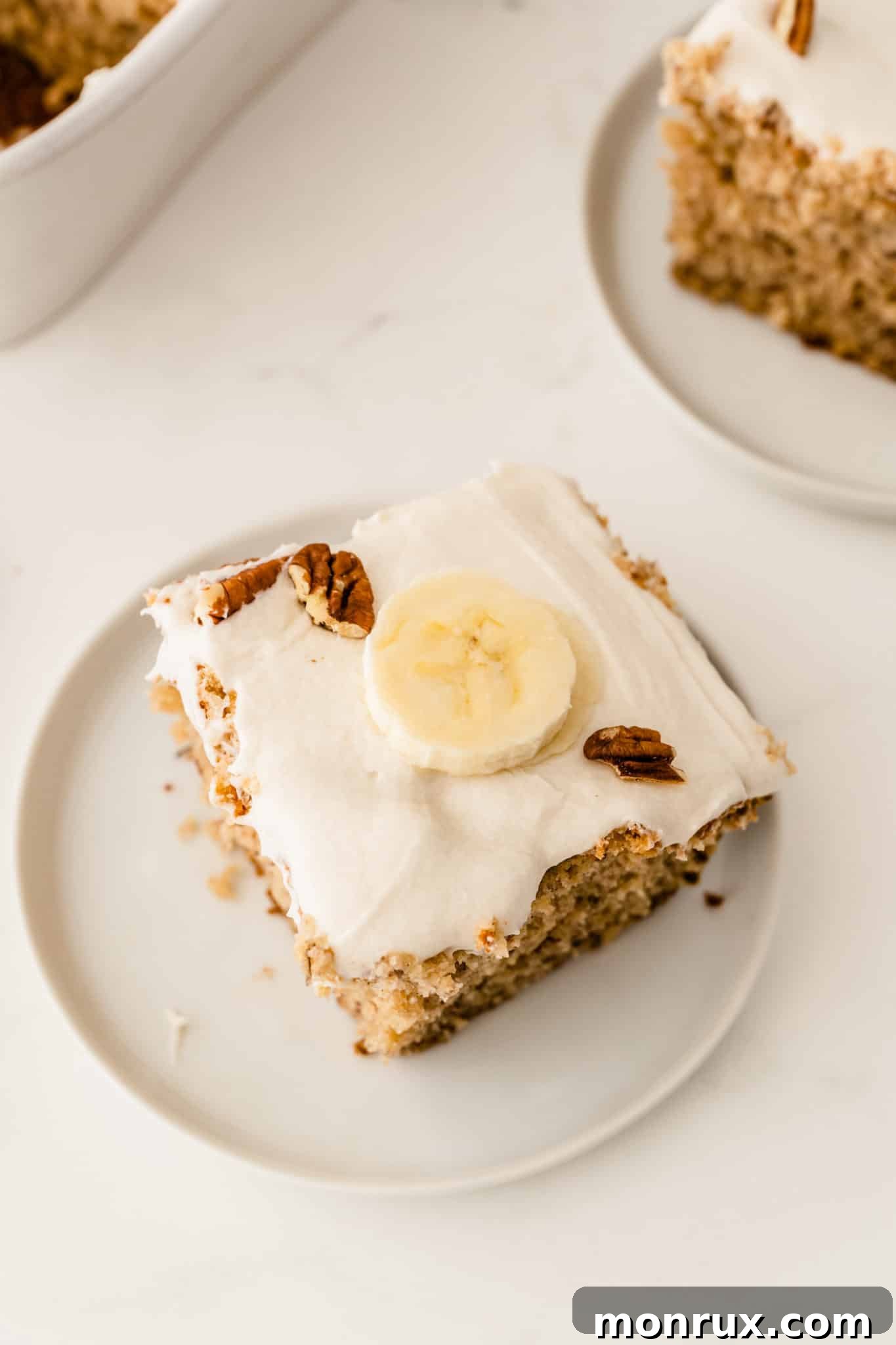 A square slice of gluten-free banana cake, frosted and garnished with a piece of banana on top.