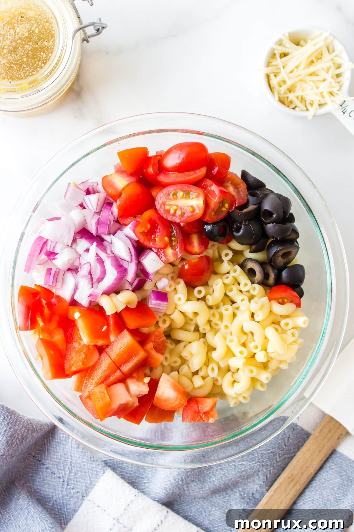 Prepared vegetables for gluten free pasta salad, including chopped red bell pepper, red onion, grape tomatoes, and black olives, gathered in a large glass mixing bowl.