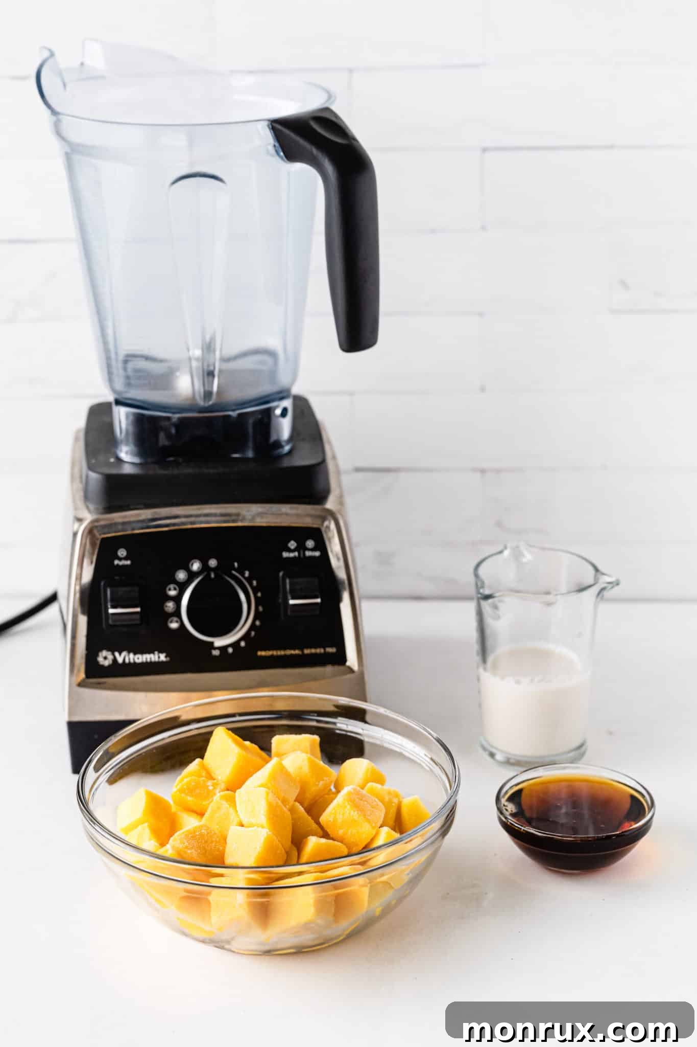 Three key ingredients for mango sorbet: frozen mango chunks, a carton of almond milk, and a bottle of maple syrup, neatly arranged on a rustic wooden table.