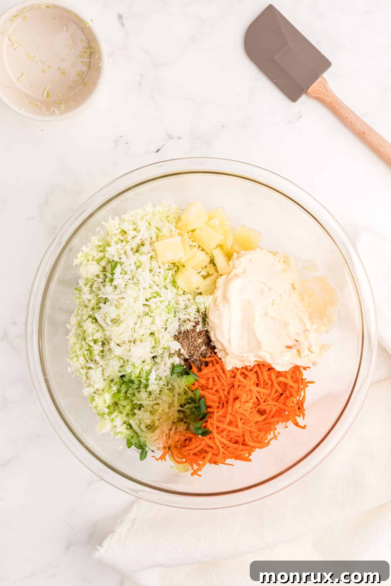 A large mixing bowl containing shredded carrots, cabbage, pineapple tidbits, and mayonnaise, ready to be combined.