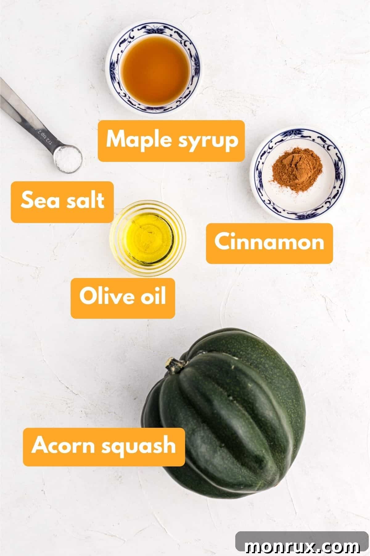 A selection of fresh ingredients including a whole acorn squash, olive oil, cinnamon, sea salt, and maple syrup, artfully arranged on a wooden surface.