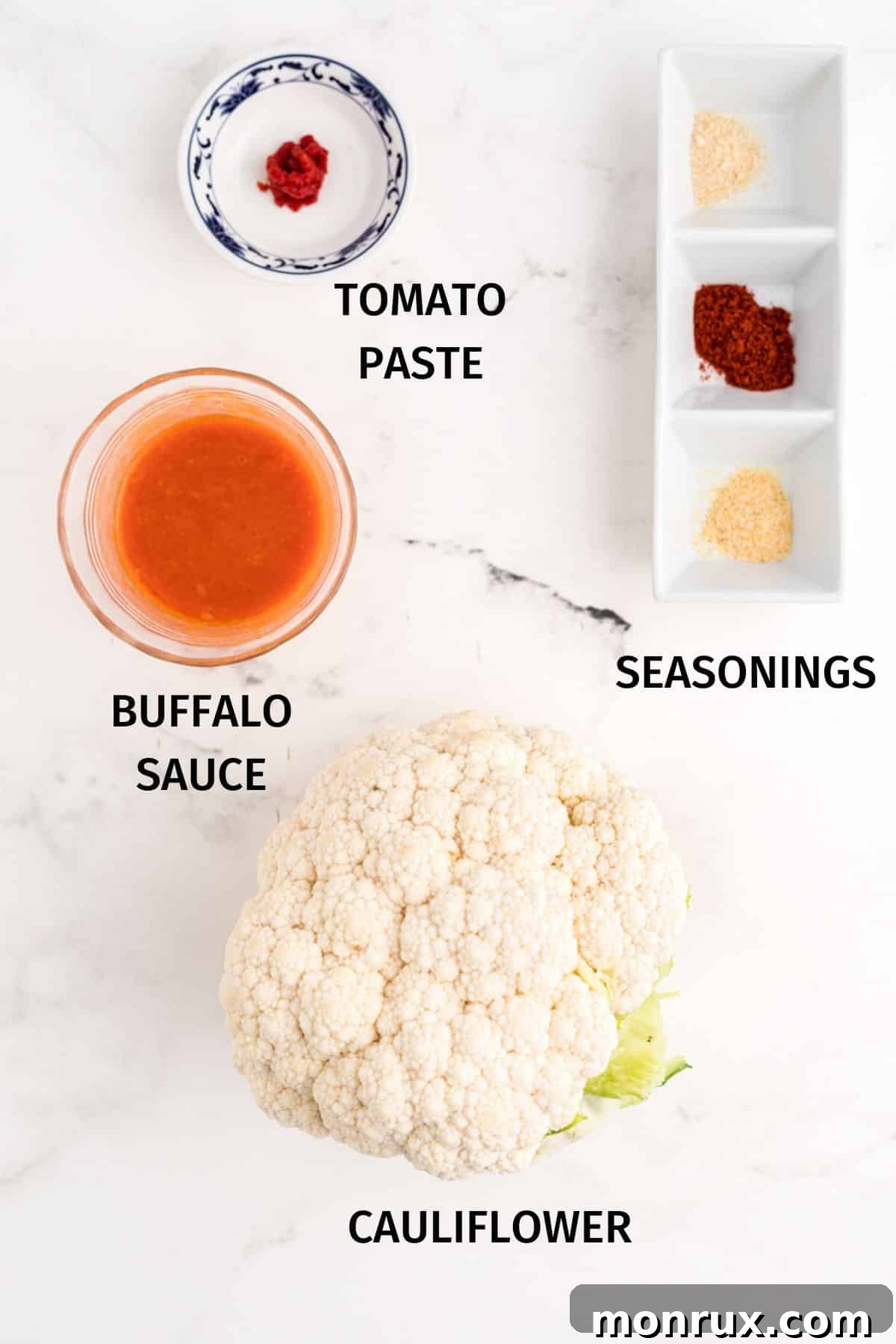An array of simple ingredients including a whole head of cauliflower, a bottle of buffalo sauce, a tube of tomato paste, and various spices, laid out on a wooden surface.