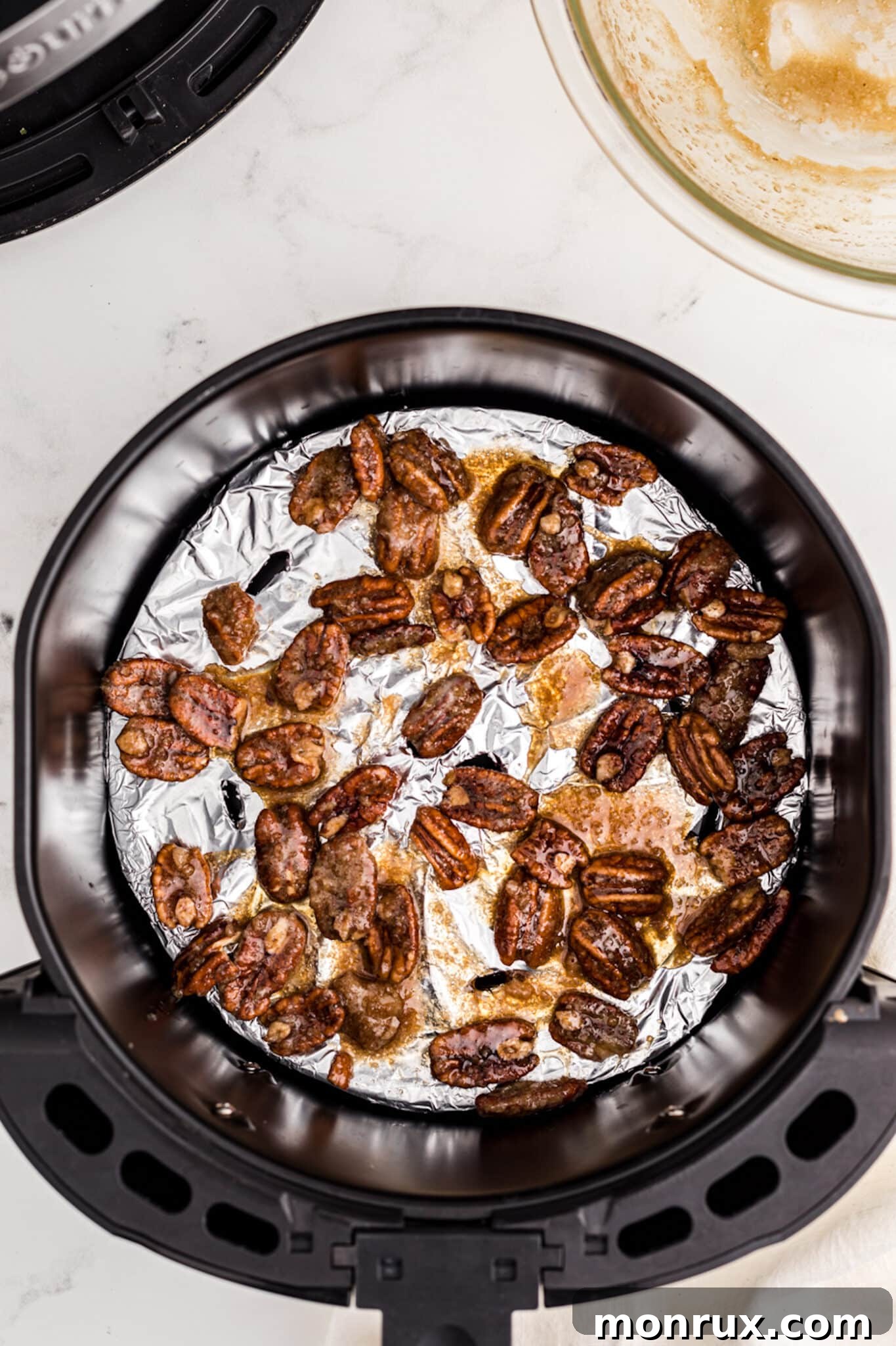 The coated candied pecans spread out in a single layer in an air fryer basket lined with parchment paper, ready for cooking.