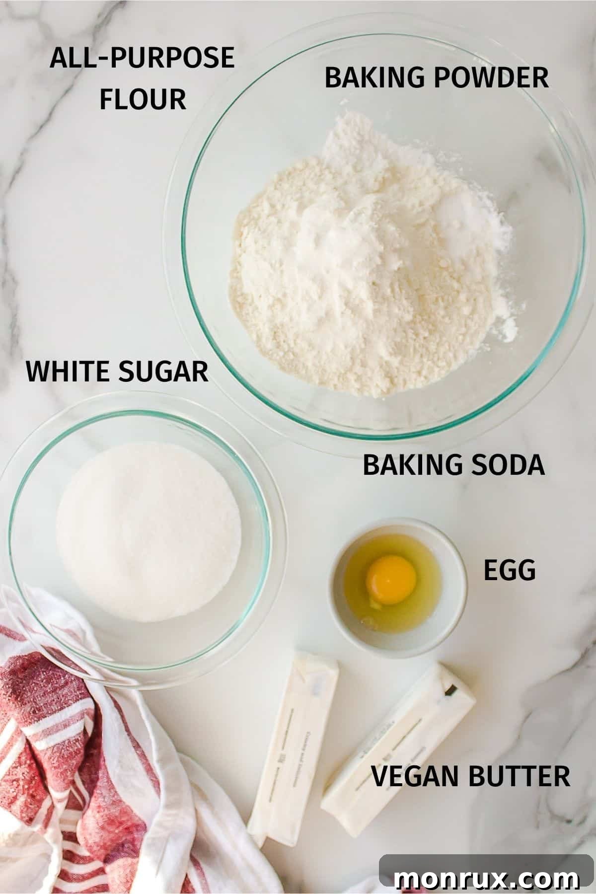 Assortment of ingredients laid out for making vegan sugar cookies, including flour, sugar, vegan butter, and an egg.