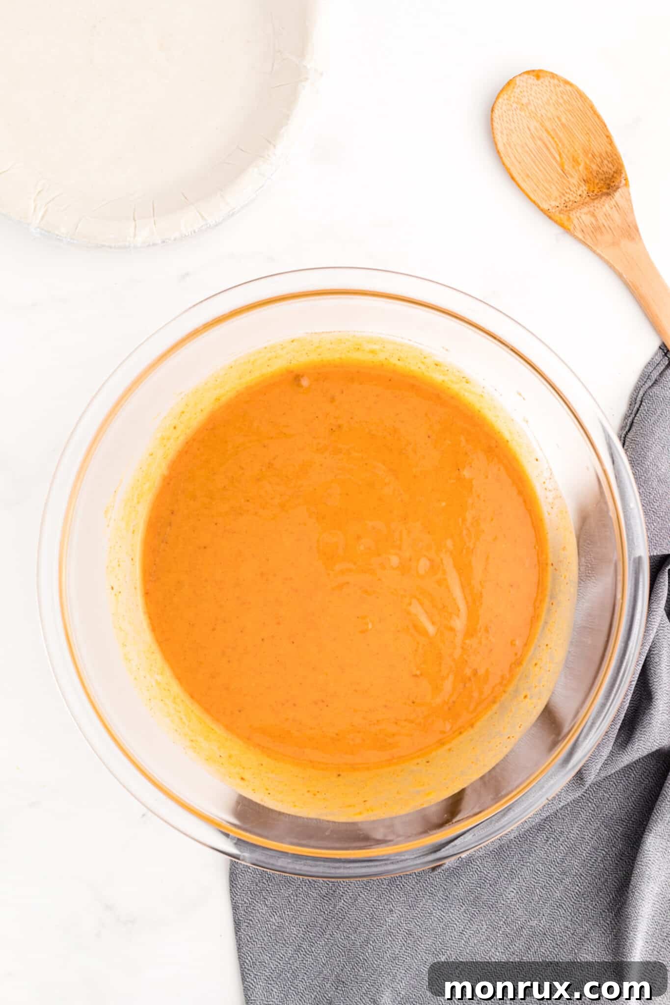 The fully mixed, smooth dairy-free pumpkin pie filling, a rich orange color, sits ready in a bowl next to an uncooked gluten-free pie crust, awaiting assembly.