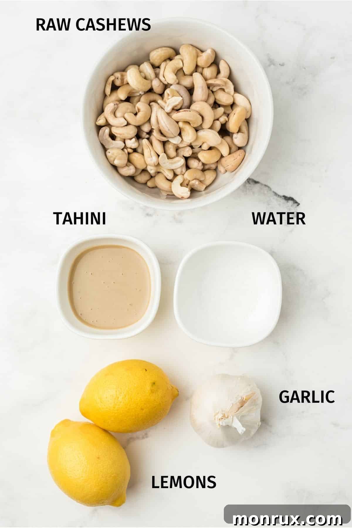 A flat lay photograph showcasing the raw ingredients for cashew hummus: raw cashews, a whole lemon, a jar of tahini, garlic cloves, and a small bowl of sea salt.