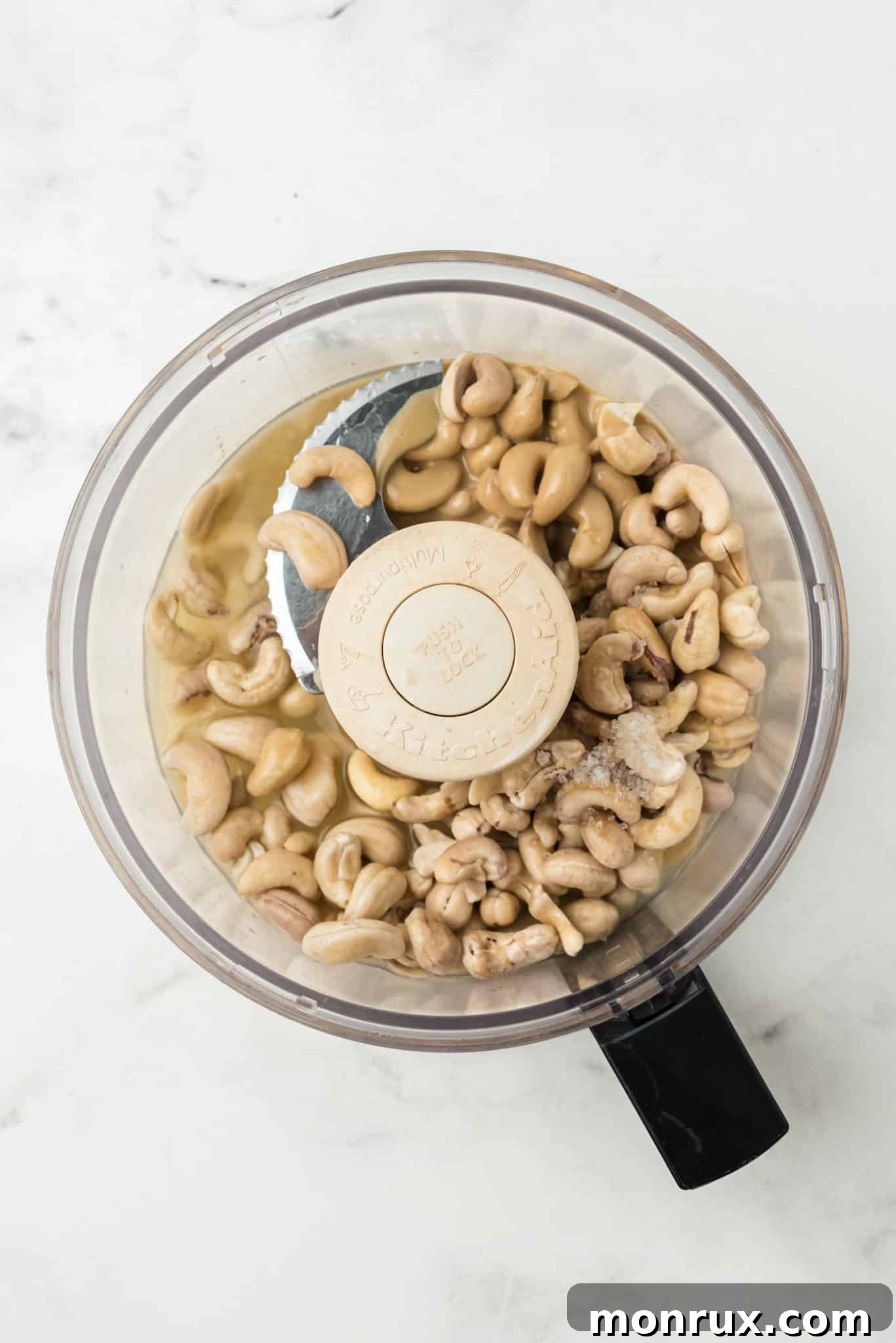 Raw cashews, lemon halves, tahini, and garlic cloves are placed inside a food processor bowl, ready to be blended into hummus.