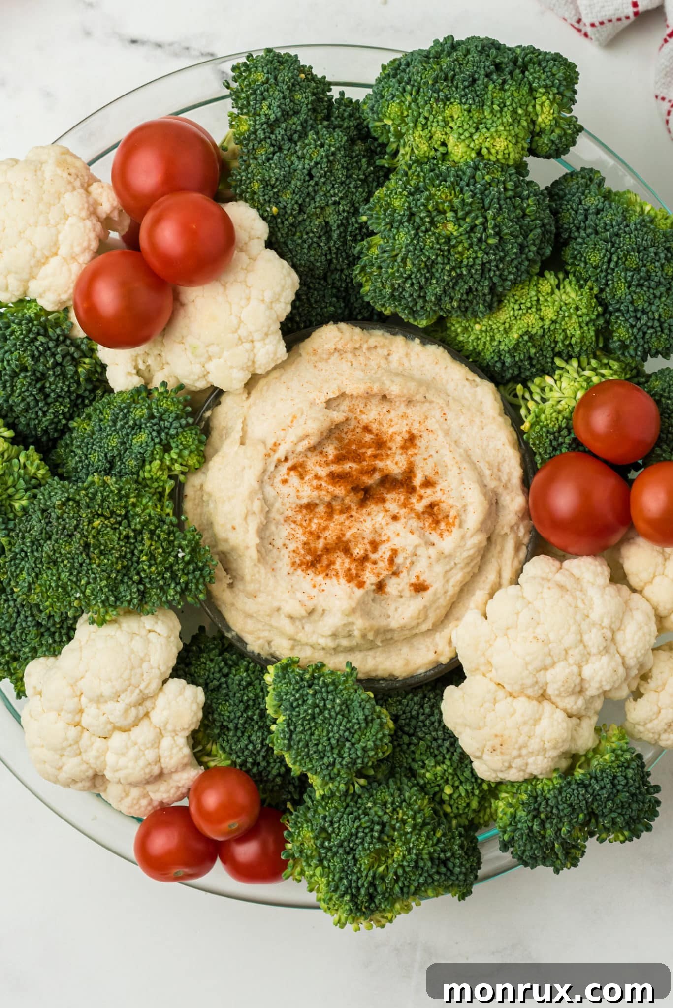 A festive holiday vegetable wreath tray with a bowl of creamy hummus in the center, adorned with fresh broccoli, cauliflower, and cherry tomatoes.