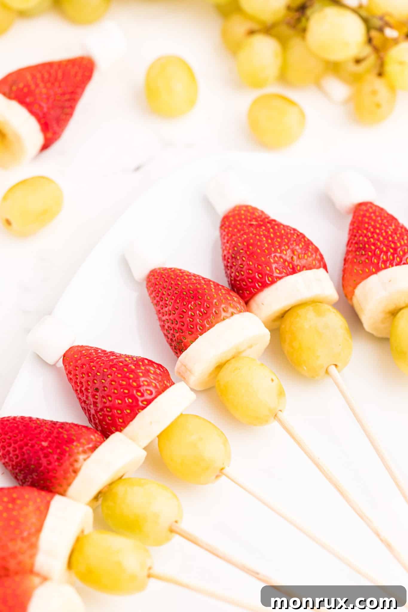 Vibrant green Grinch fruit kabobs with grapes, bananas, strawberries, and marshmallows artfully arranged on a white plate, ready for a healthy holiday snack.
