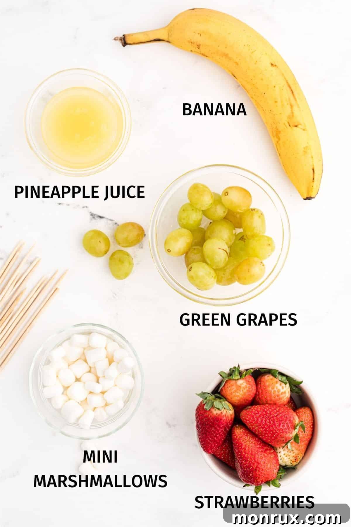 A flat lay photograph displaying the fresh ingredients required for Grinch fruit kabobs: green grapes, sliced bananas soaking in pineapple juice, vibrant red strawberries, and a small bowl of mini marshmallows.
