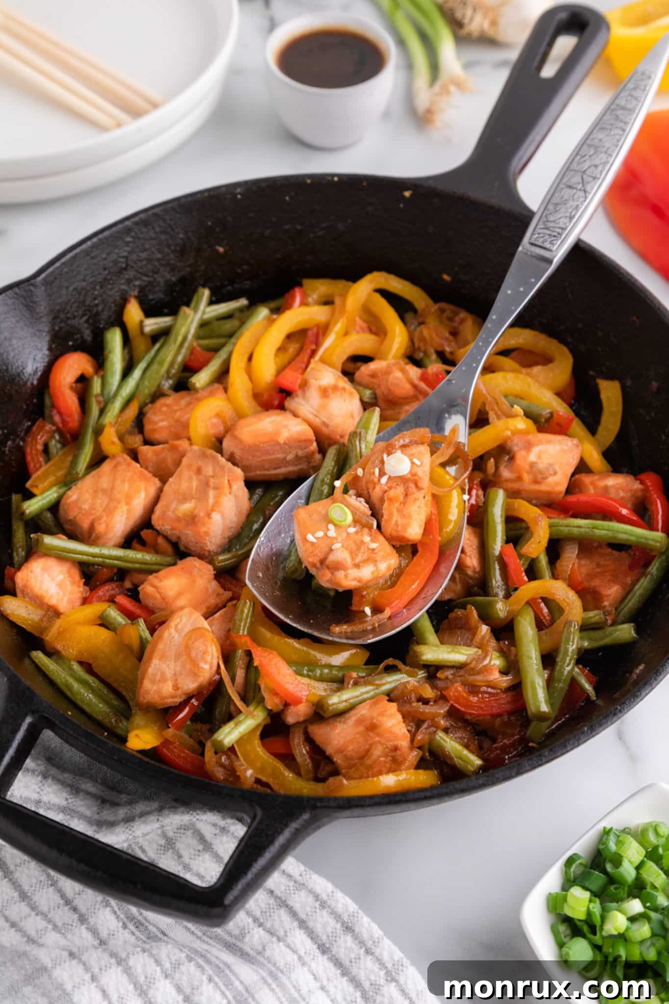 A silver serving spoon lifting a portion of salmon stir-fry from a rustic cast iron skillet, showcasing the perfectly cooked fish and vegetables.