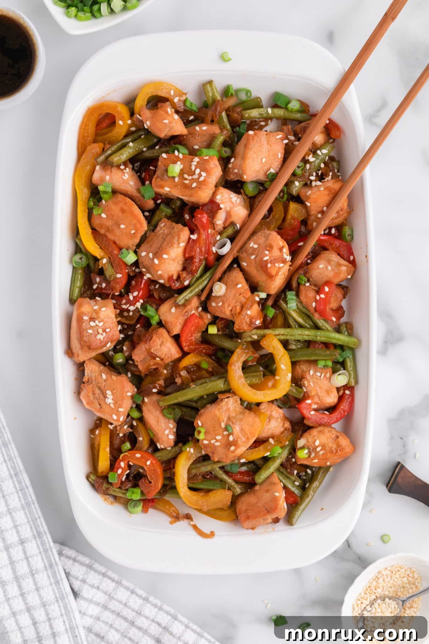 An elegantly plated dish of salmon and vegetable stir-fry, garnished with sesame seeds and scallions, accompanied by chopsticks, ready to be enjoyed.