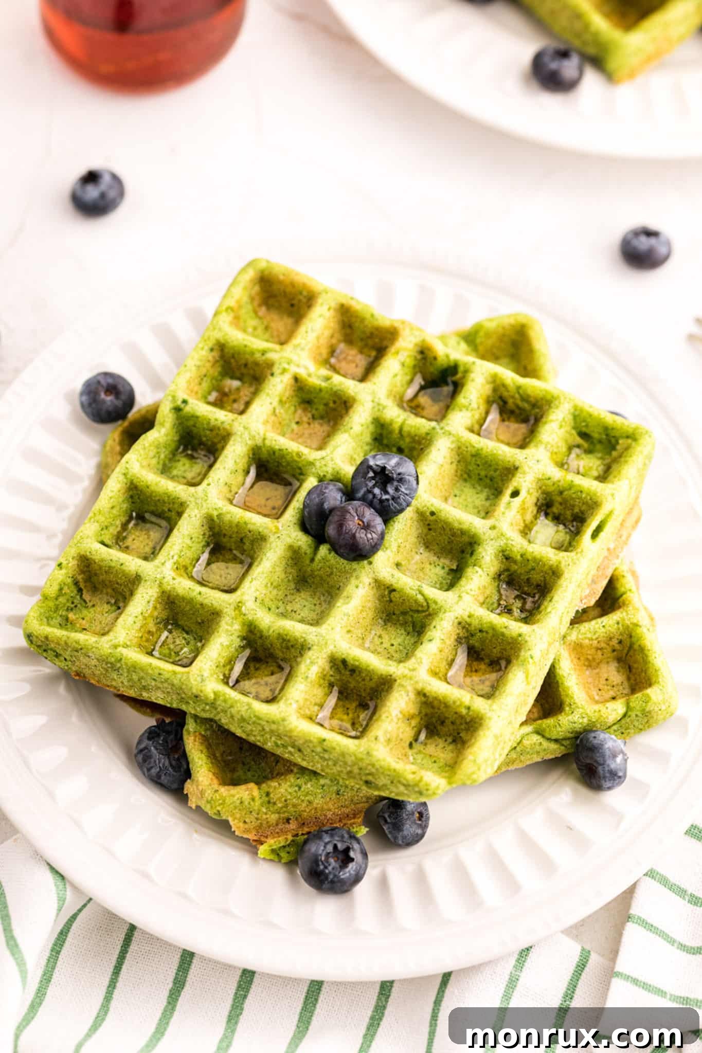 A stack of two perfectly cooked green waffles topped with fresh blueberries and a drizzle of maple syrup.