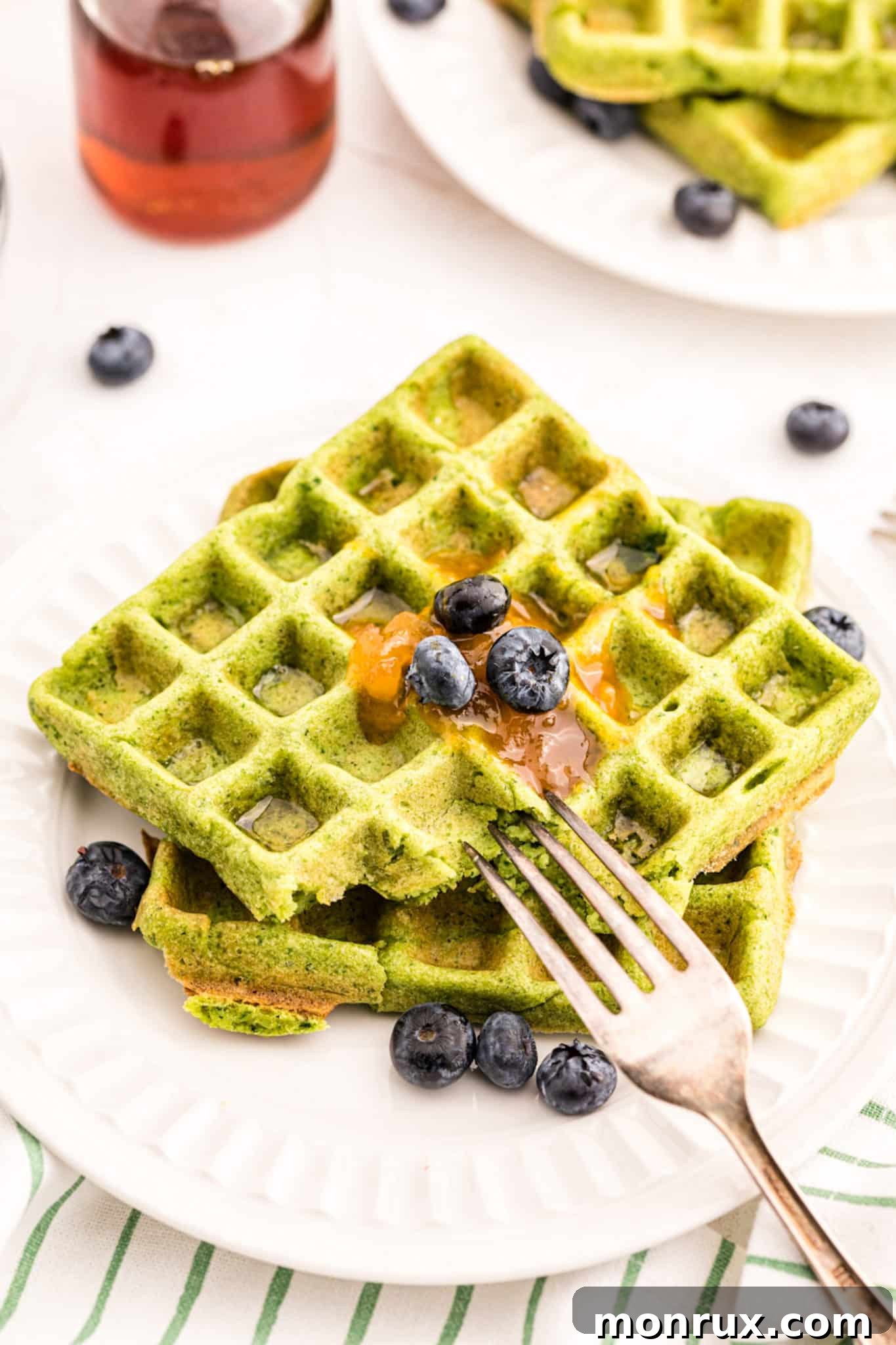 A beautifully arranged stack of green waffles on a white plate, garnished with fresh blueberries and a generous pour of maple syrup.