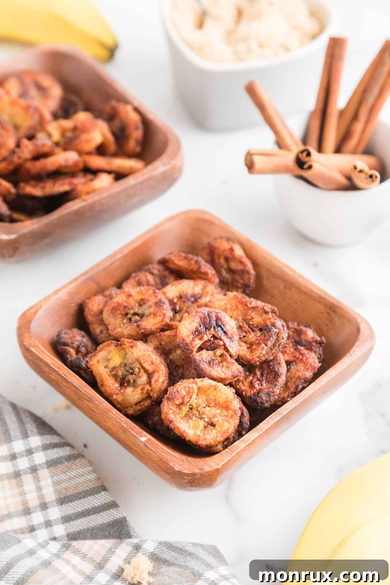 A square wooden bowl filled with air fryer cinnamon banana chips, garnished with a cinnamon stick and fresh banana slices.