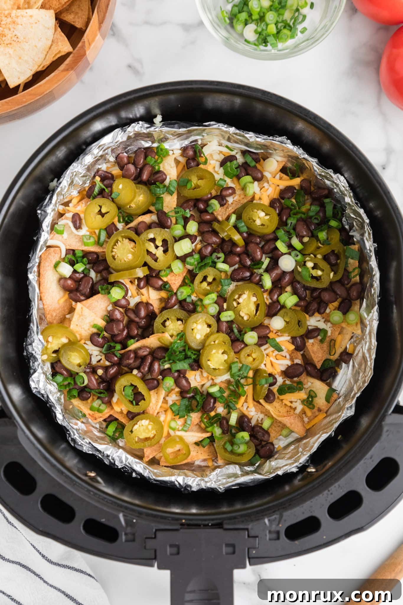 Nacho toppings of black beans and jalapenos on top of chips in an air fryer basket, ready for the final cheese layer.